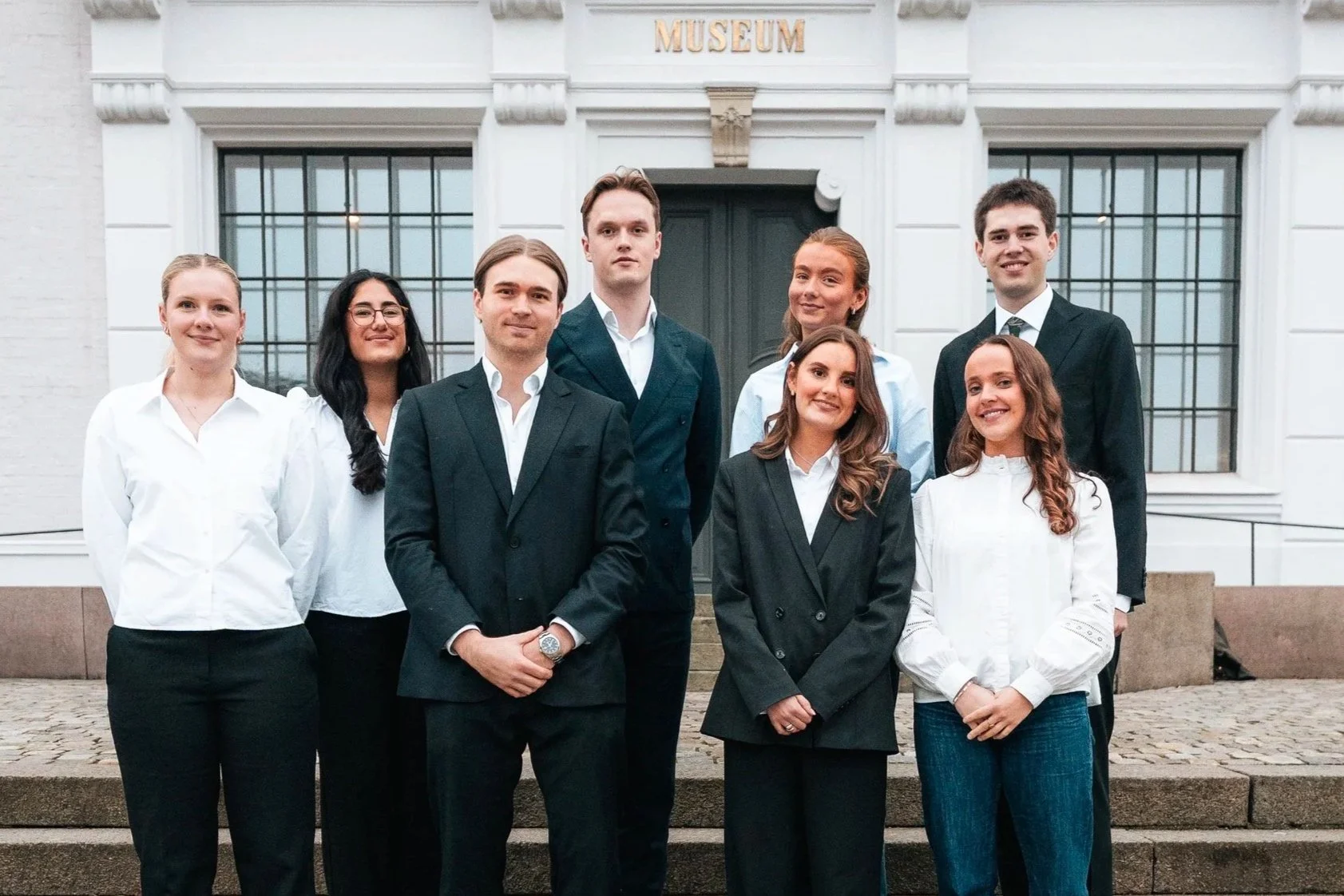 Bilde av fag- og bedriftskomiteen i Econos: en gruppe på åtte unge studenter, tre menn og fem kvinner. De står på en trapp utenfor en stor, hvit bygning med store vinduer og en mørkegrønn dør, kledd i profesjonelle og formelle antrekk.