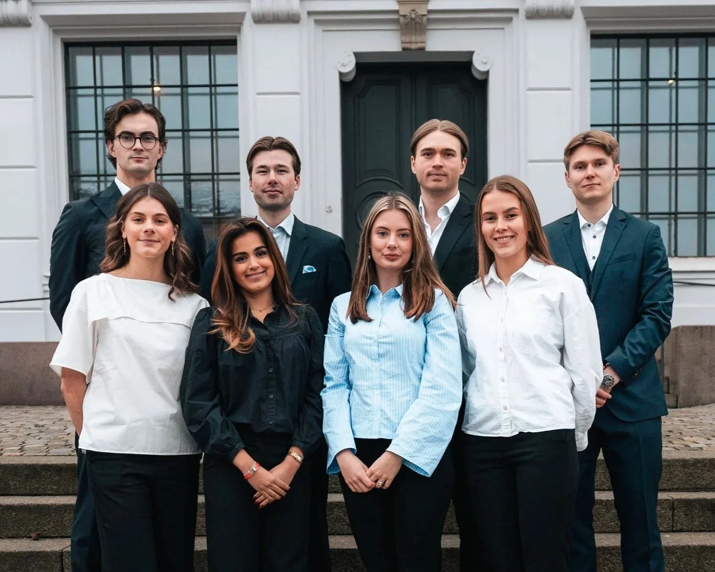 Bilde av styret i Econos: en gruppe på åtte unge studenter. De står på en trapp utenfor en stor, hvit bygning med store vinduer og en mørkegrønn dør, kledd i profesjonelle og formelle antrekk.