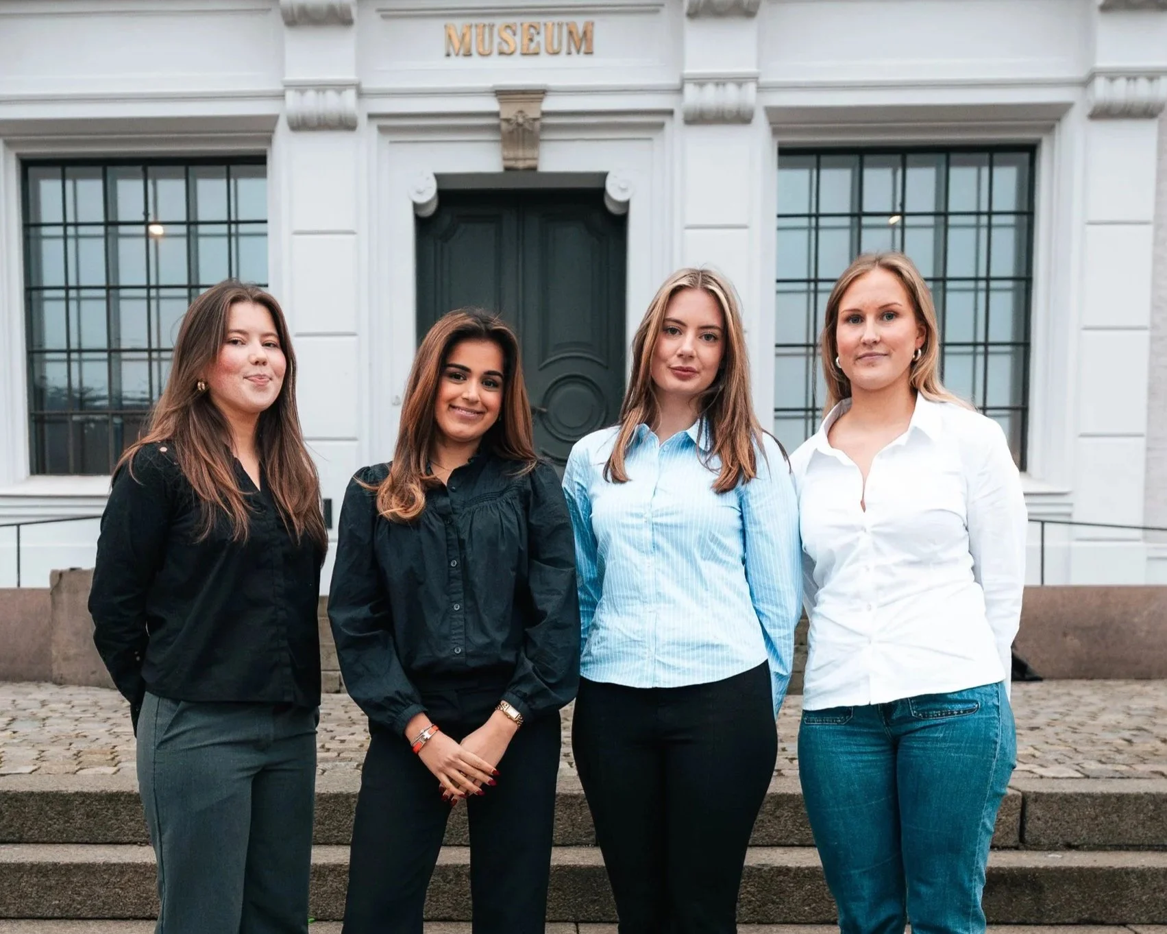 Bilde av sosialkomiteen i Econos: en gruppe på fire unge studenter. De står på en trapp utenfor en stor, hvit bygning med store vinduer og en mørkegrønn dør, kledd i profesjonelle og formelle antrekk.