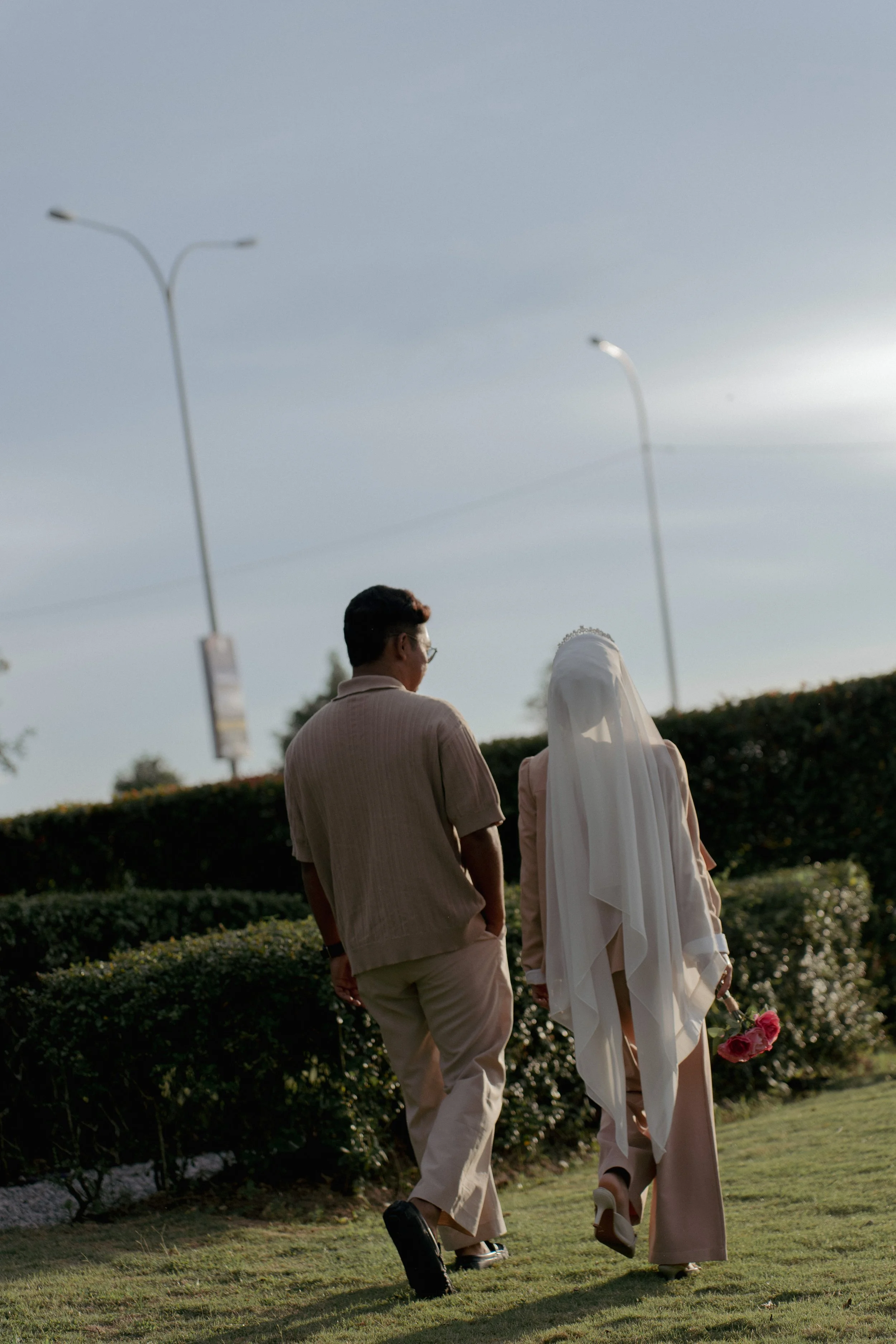 A woman dressed in a white wedding gown and veil holding pink roses walking with a man in a beige outfit on a grassy area outdoors.