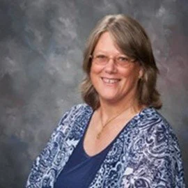 A man with curly dark hair, light skin, wearing clear glasses and a nose ring, looking to the side with a slight smile, in a room with a white wall and a green plant in the background.