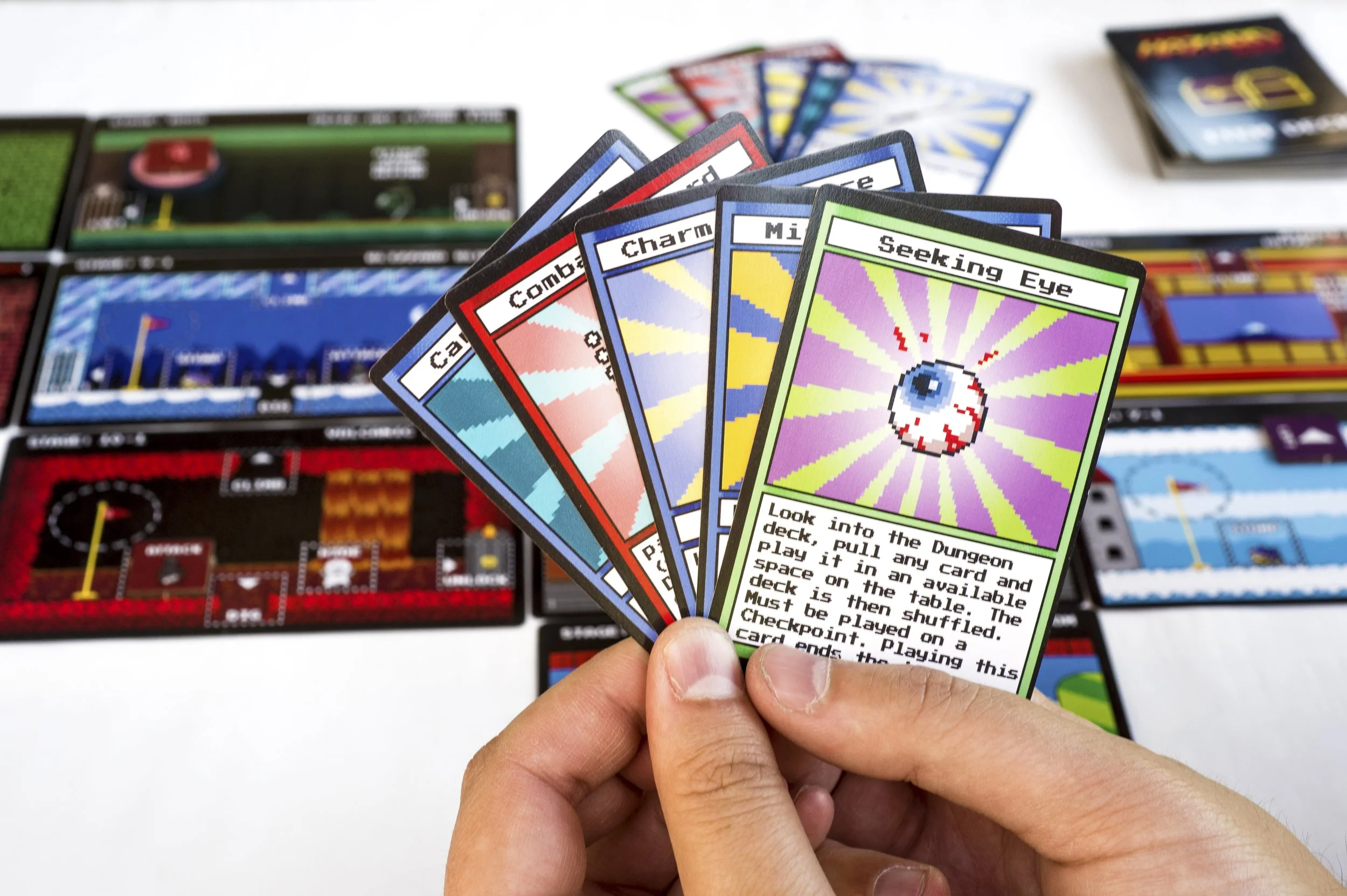 Close-up of a hand holding five colorful pixelated cards from a video game card game, with game boards and cards in the background.