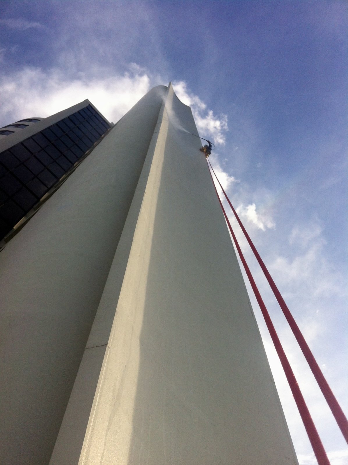 A worker in safety gear cleaning the tall exterior wall of a skyscraper with a red rope hanging from the top, view from the ground looking up.