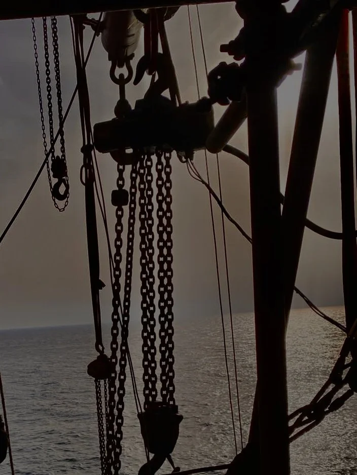 A silhouette of rigging equipment with chains and pulleys against the ocean and sunset background.