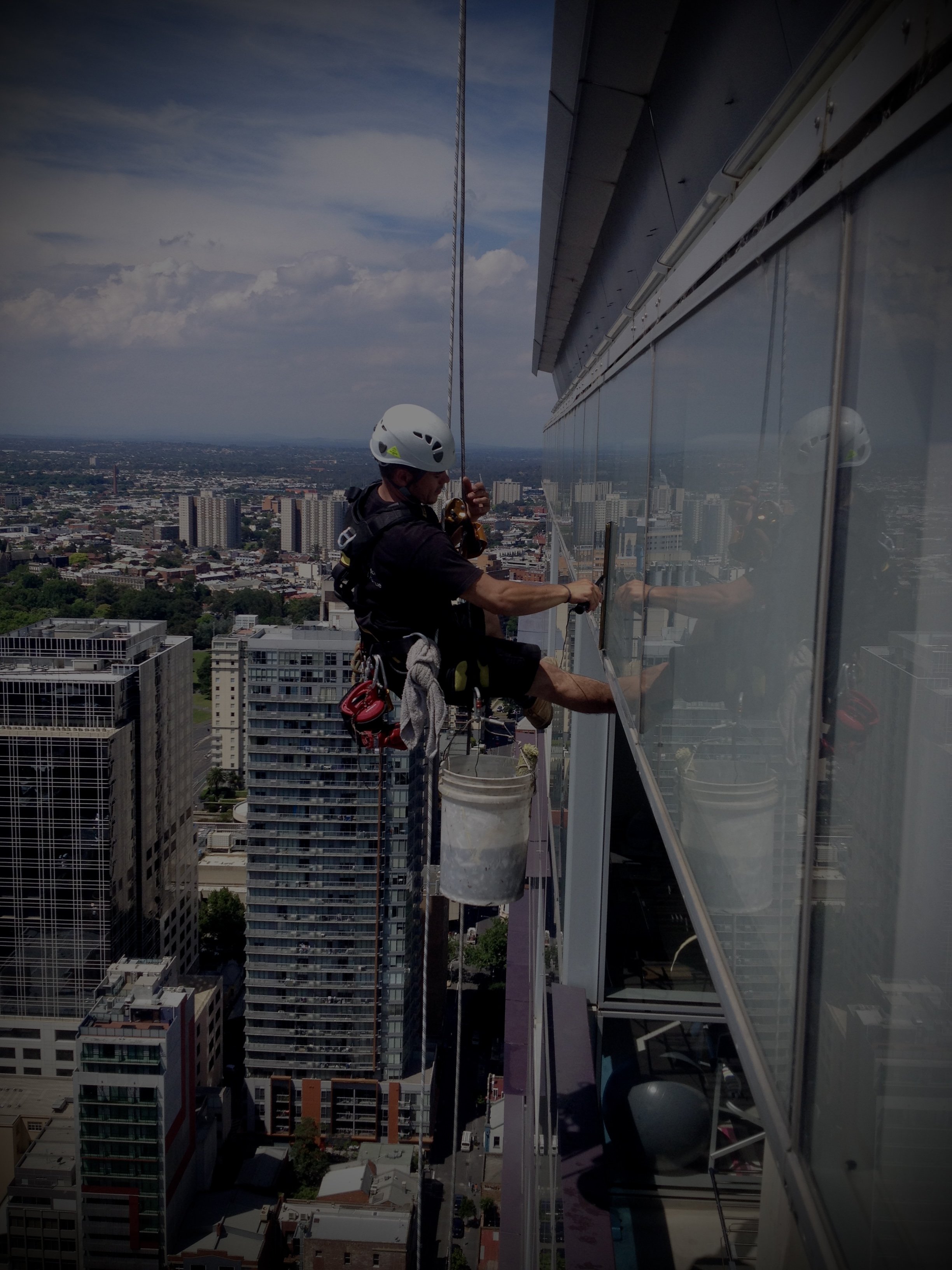 A window cleaner wearing a safety helmet and harness cleaning the exterior glass wall of a high-rise building in a city.