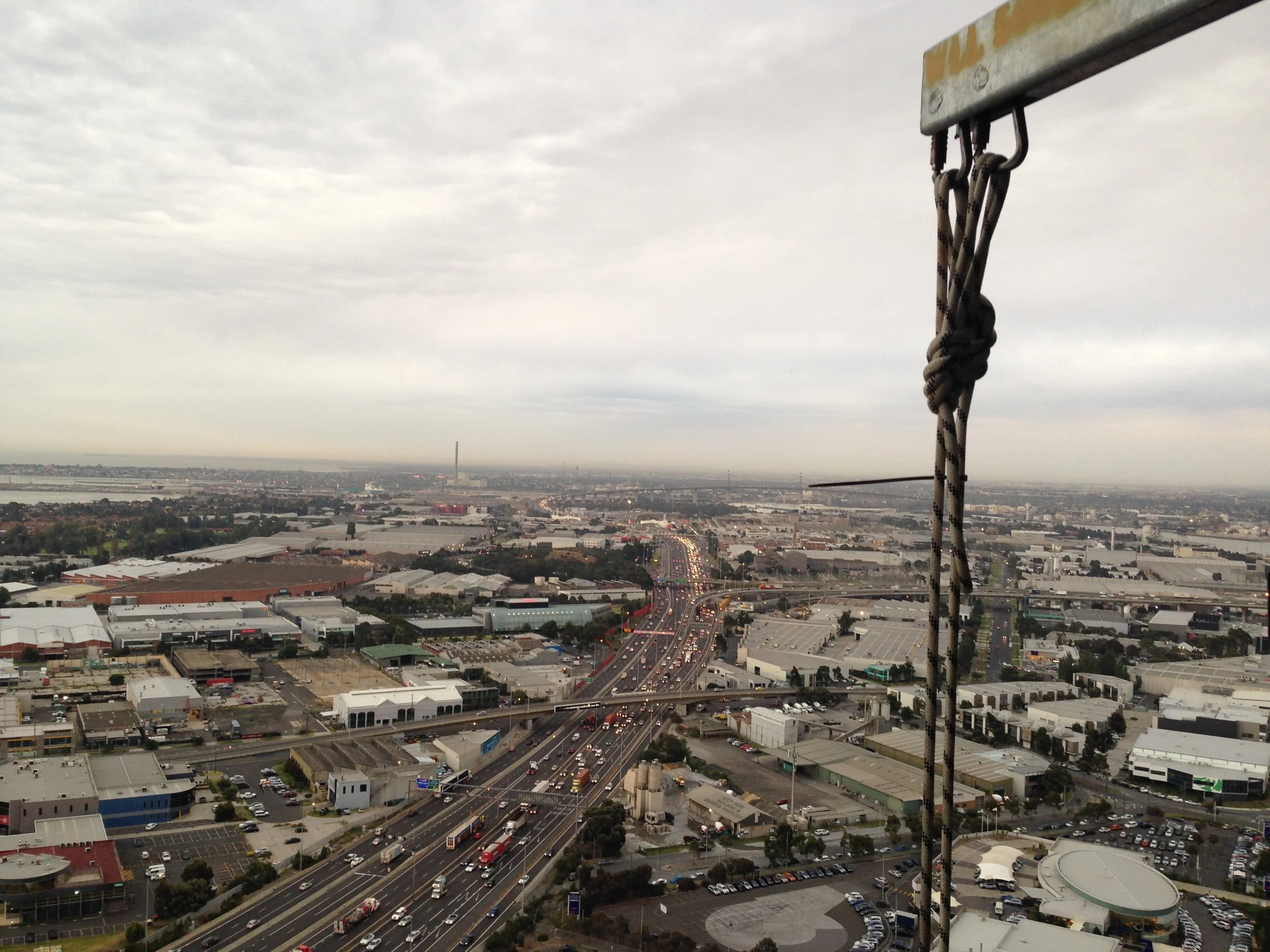 A cityscape view from a high vantage point showing rope access rope, highways, buildings, and industrial areas under a cloudy sky.