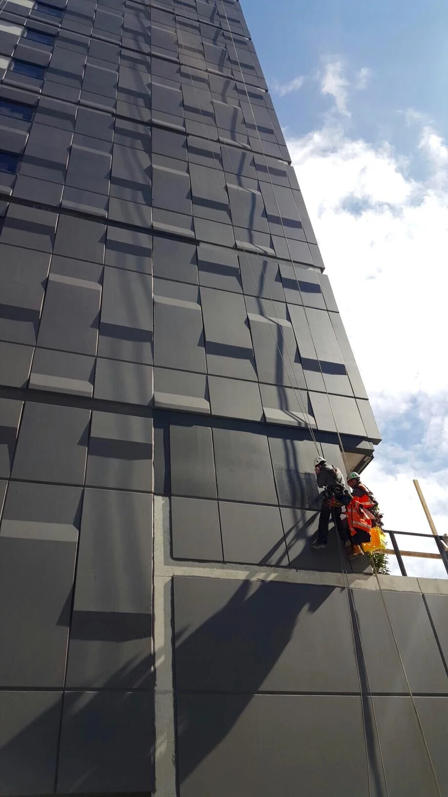 Two rope access technicians inspecting and painting the exterior of a tall modern building with dark panels under a partly cloudy sky.
