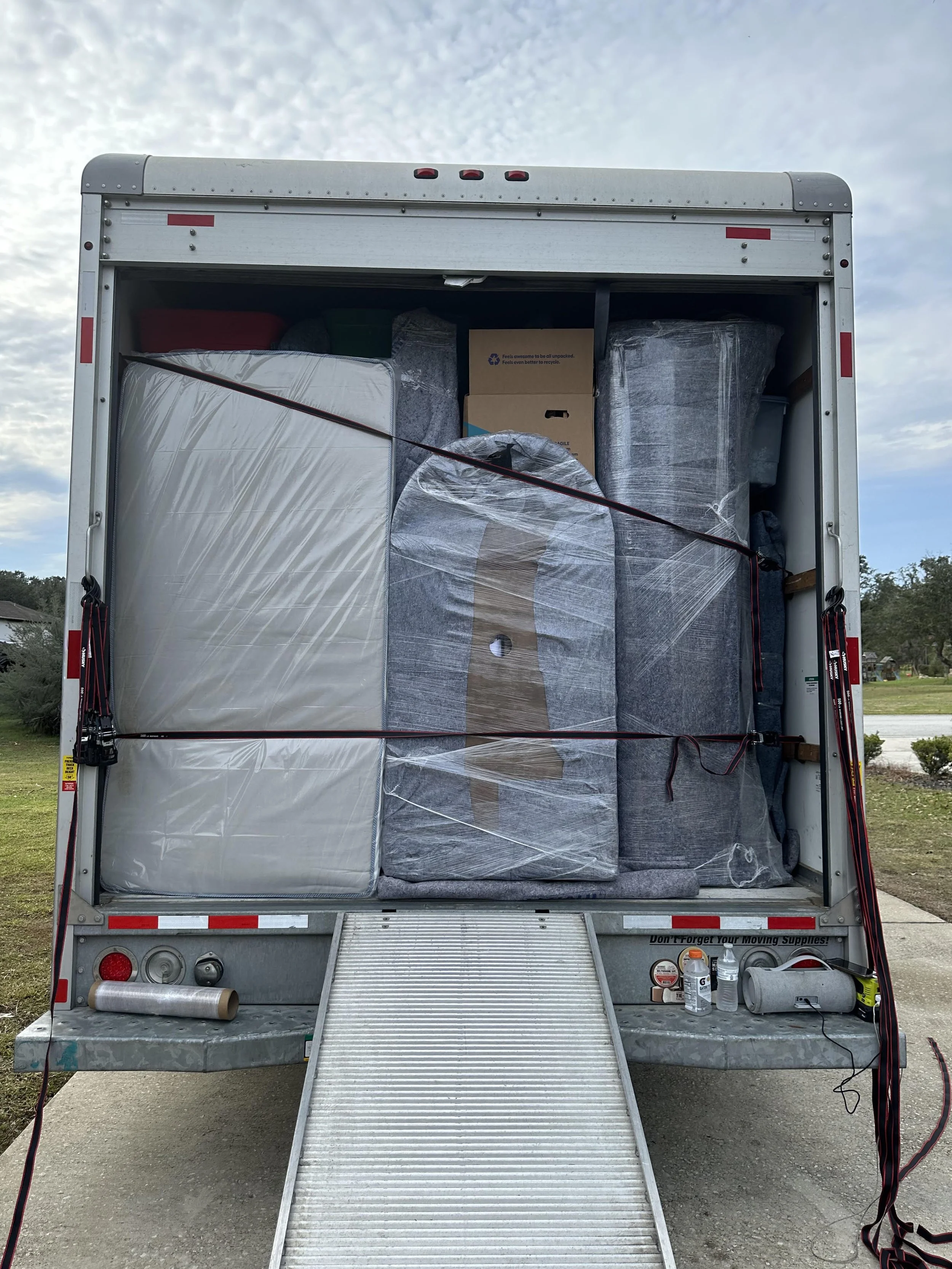 Back of a moving truck filled with wrapped furniture and boxes, secured with straps, parked on a driveway with a cloudy sky and trees in the background by Easy Peasy Moving, LLC, a moving company in Altamonte Springs and surrounding areas of Florida.