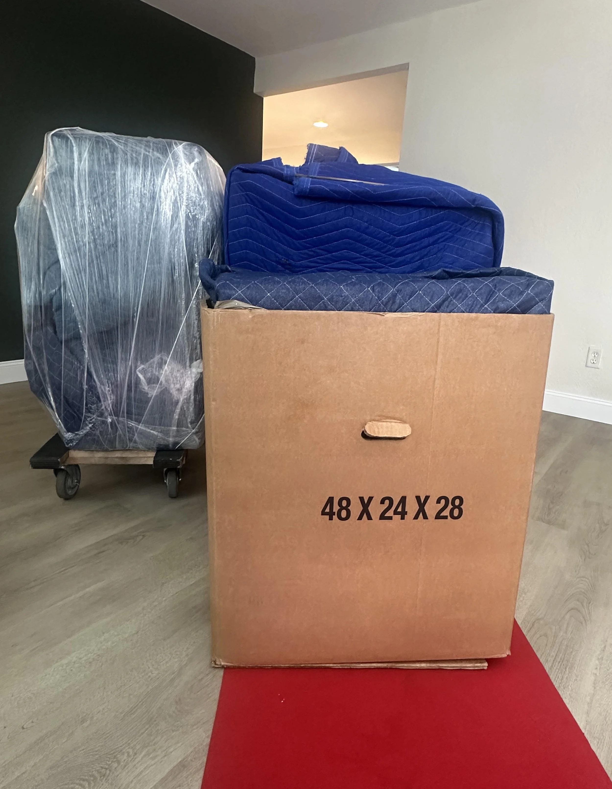 A large cardboard box with size dimensions 48 x 24 x 28 printed on it, filled with stacked blue mattresses, and some mattresses wrapped in plastic next to it, on a red mat on a wooden floor inside a room by the moving company, Easy Peasy Moving, LLC.