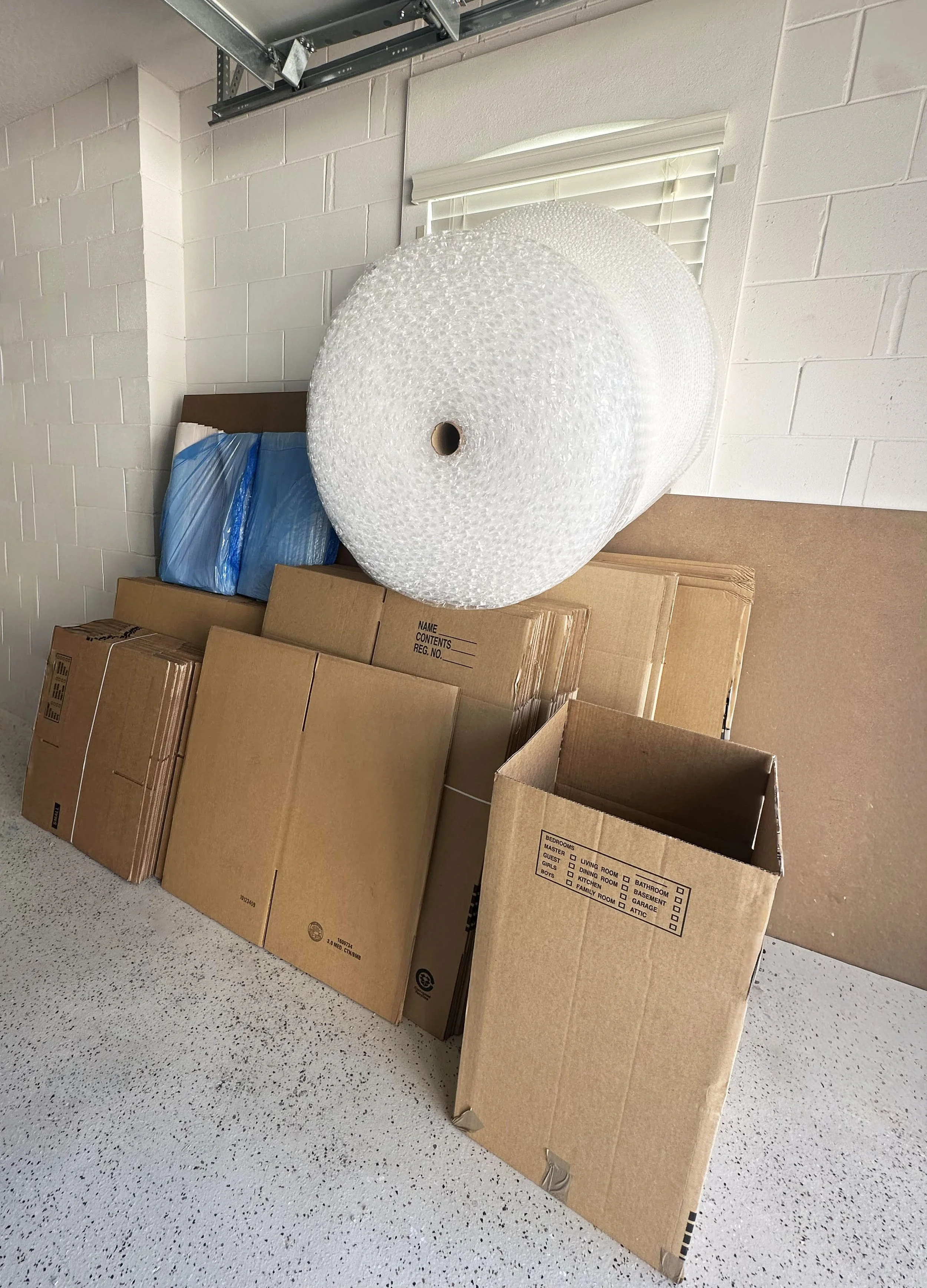 A corner of a room with stacked cardboard boxes, some with packing labels, and two large rolls of bubble wrap leaning against the wall near a window with blinds for a move by Easy Peasy Moving, LLC in Central Florida.