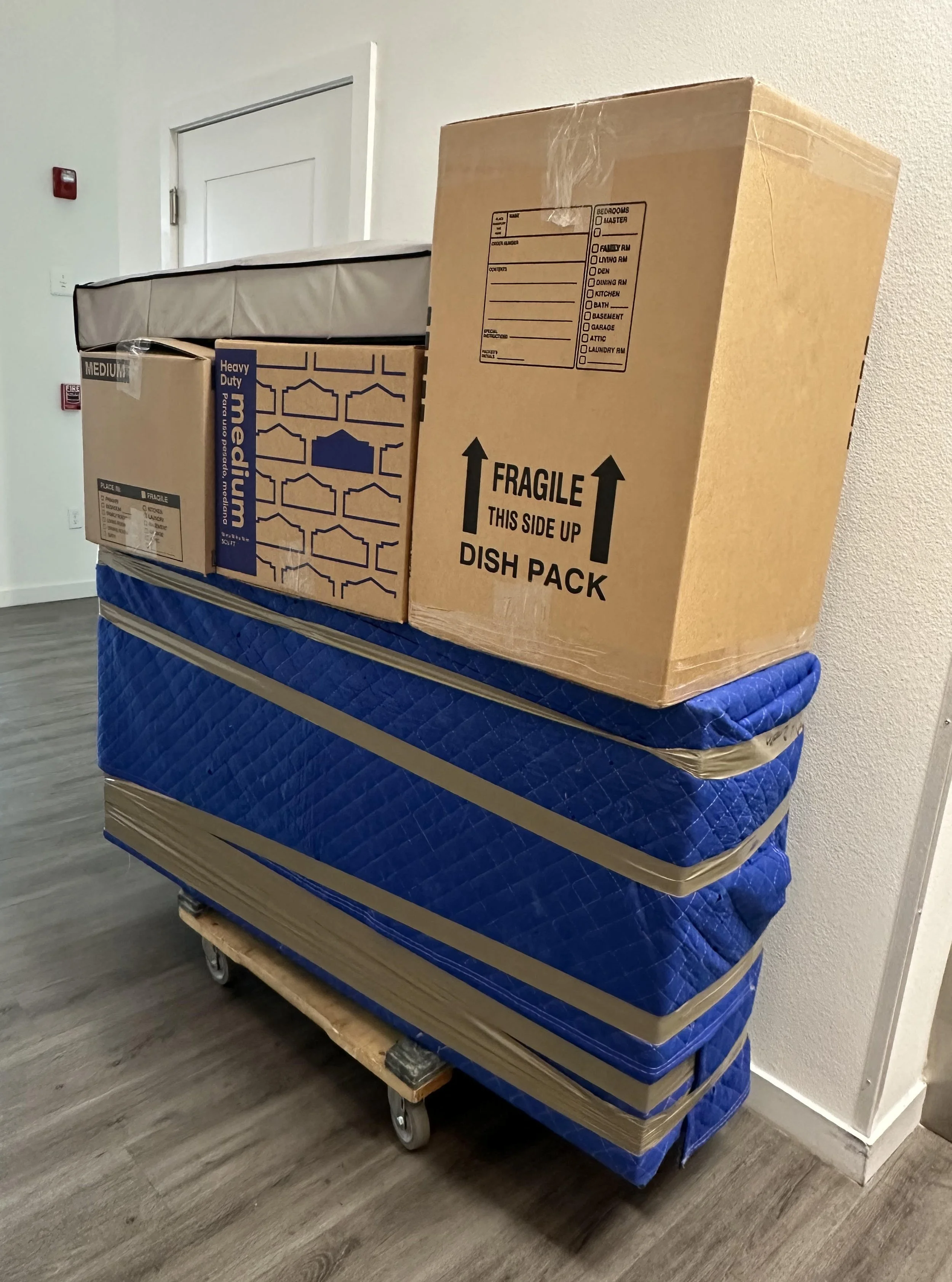 Stack of moving boxes and a mattress on a dolly against a white wall in a hallway by Easy Peasy Moving, LLC, a moving company in the Altamonte Springs area of Florida.