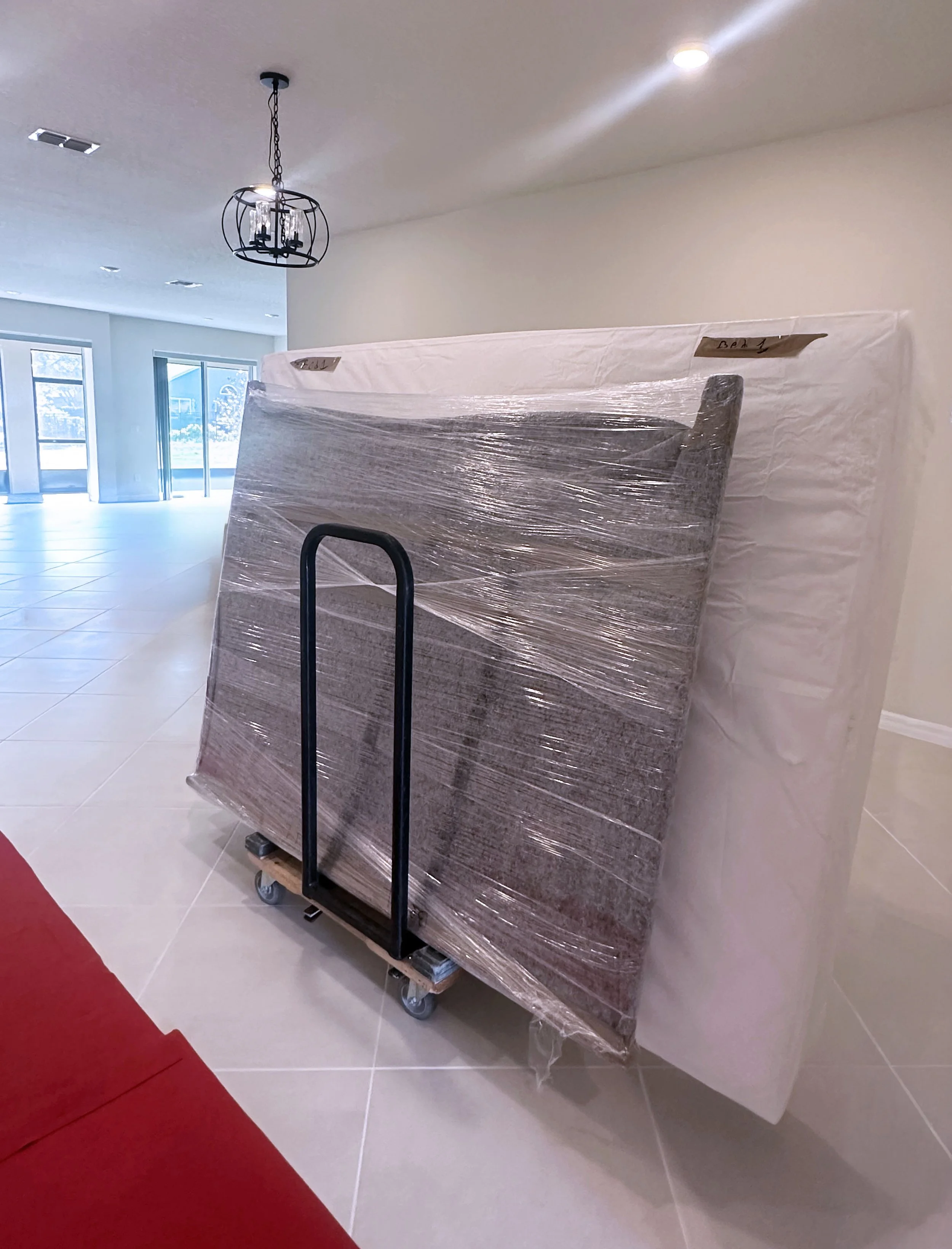 Large furniture piece wrapped in plastic on a dolly in a spacious, well-lit room with large sliding glass doors and a chandelier in Central Florida, photographed by Easy Peasy Moving, LLC crew member.