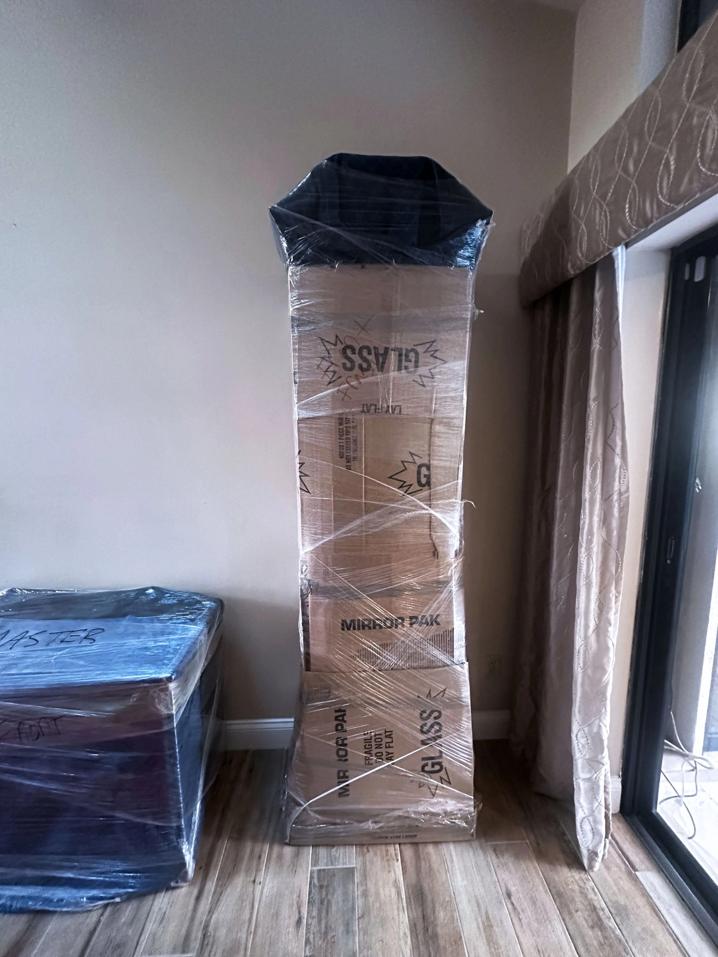 Stack of boxes wrapped in plastic wrap in a room with wooden floors, next to a sliding glass door with curtains by the moving company, Easy Peasy Moving, LLC in Central Florida.