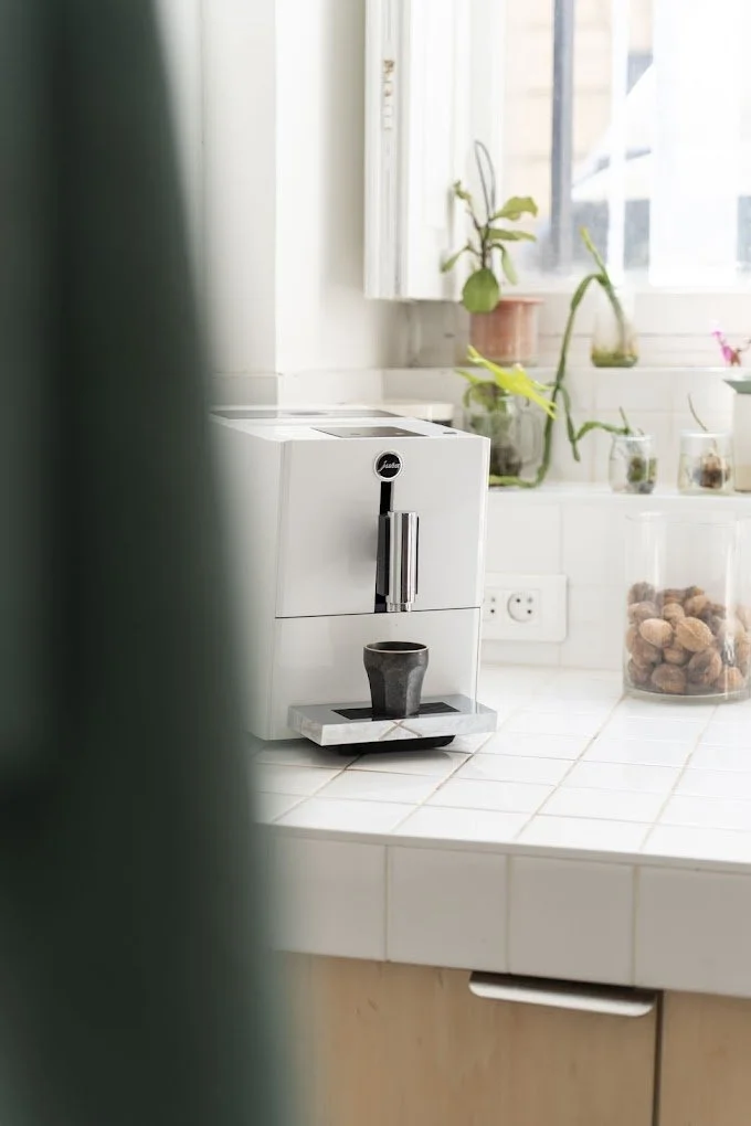 Une machine à café blanche sur un plan de travail de cuisine blanc avec une tasse en dessous, des plantes en pot sur la fenêtre en arrière-plan et un grand bocal transparent rempli de noix.