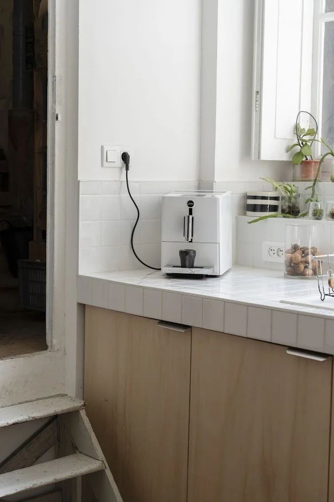Une machine à café expresso blanche sur un comptoir de cuisine avec des plantes, une prise électrique, et un miroir en arrière-plan.