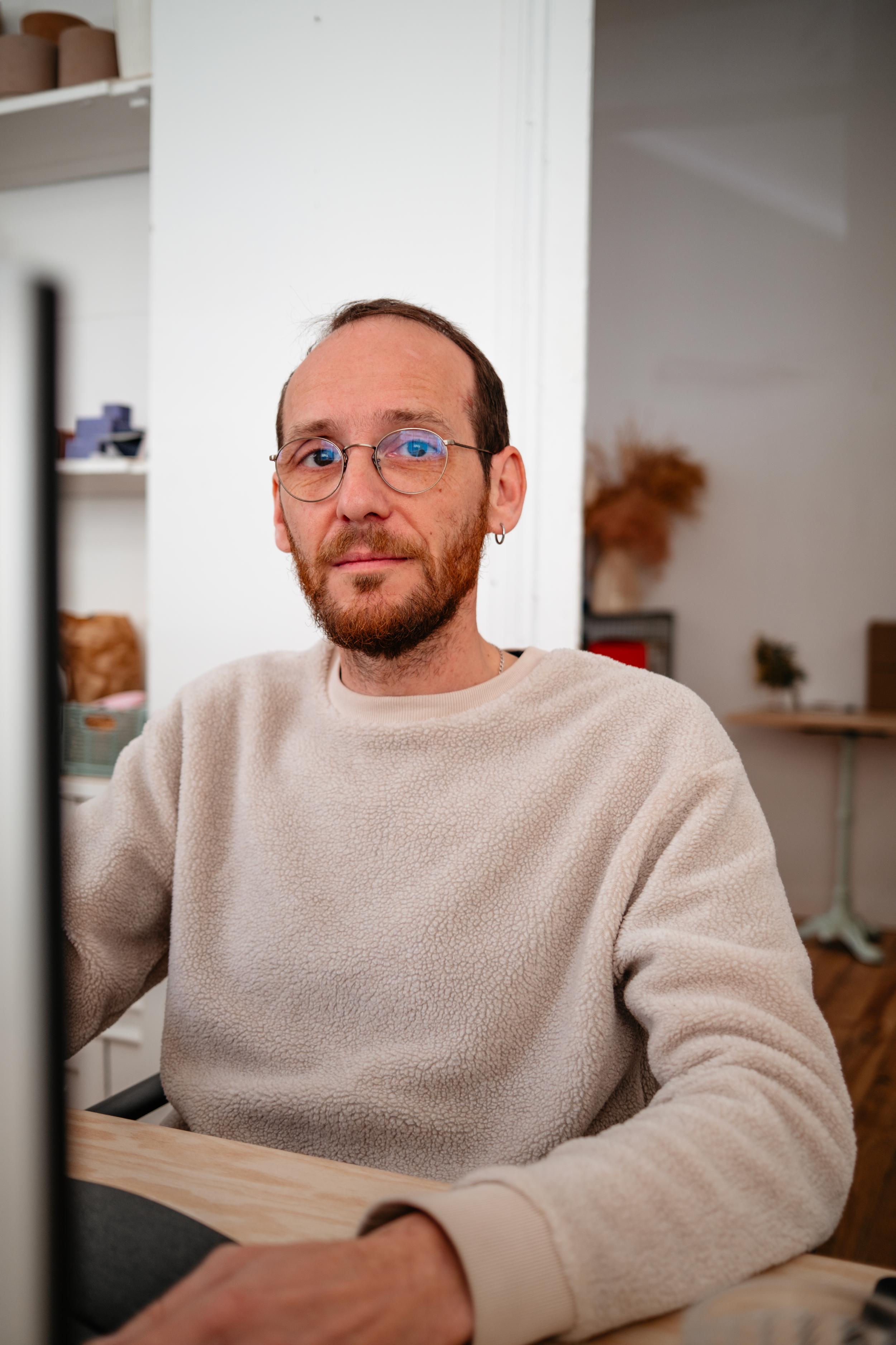 Homme portant un chapeau noir, lunettes noires, assis à une table en bois, travaillant sur un ordinateur portatif, avec une vue sur une forêt par une grande fenêtre.