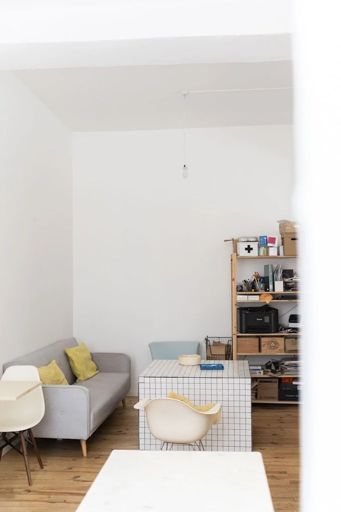 Salon avec un canapé gris, une table à carreaux et des chaises, étagère en bois avec livres et accessoires, sol en bois, mur blanc, lumière suspendue