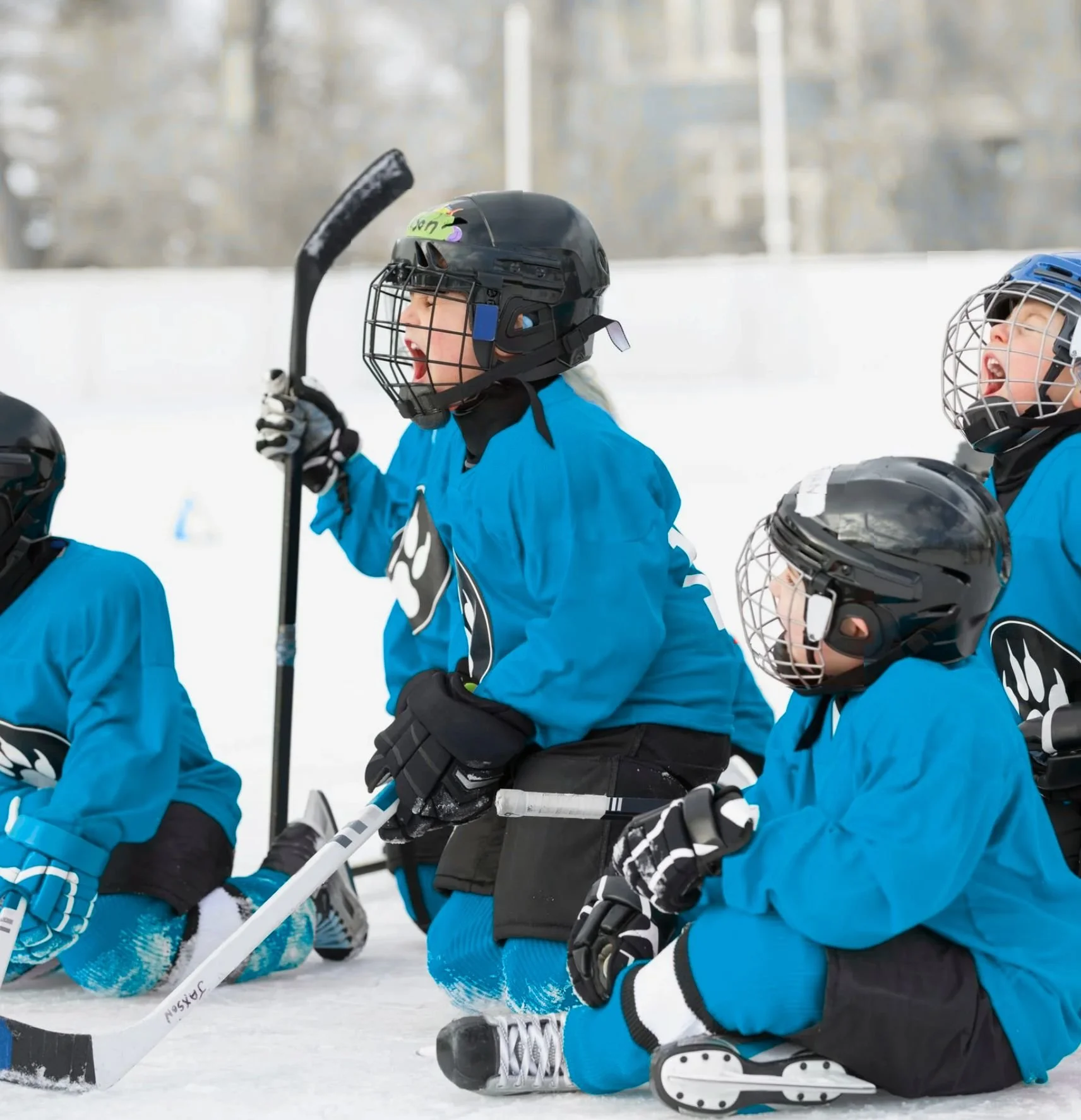 Children in blue hockey jerseys and helmets kneeling on an ice rink, holding hockey sticks and cheering during a game or practice.