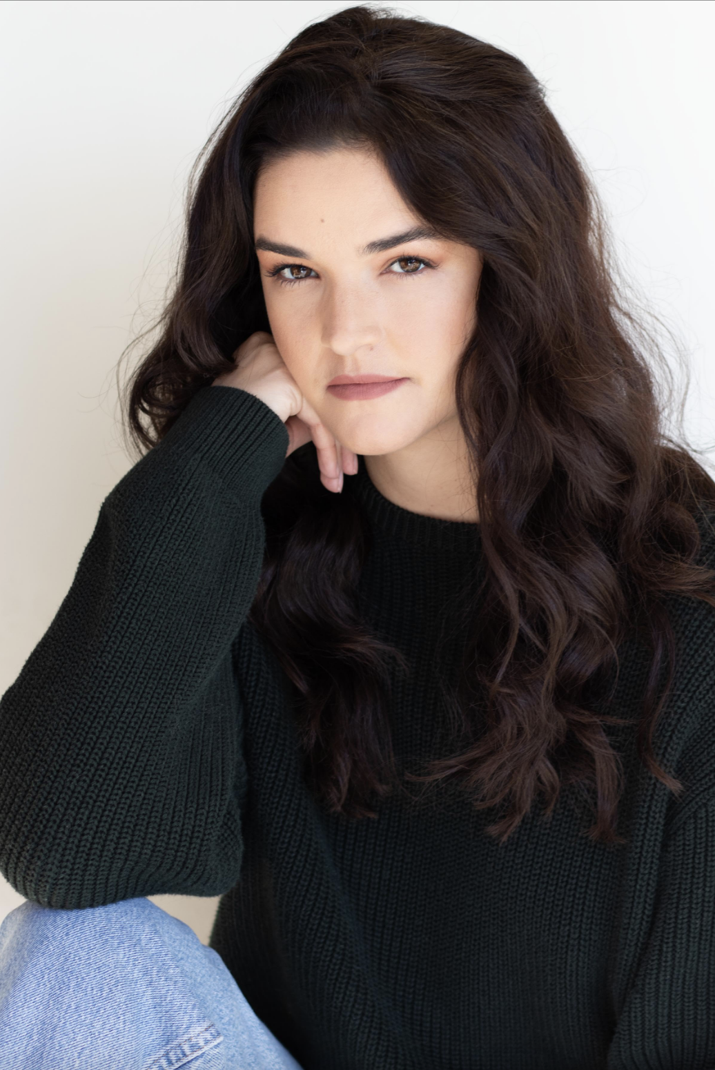 Close-up portrait of a woman with long, wavy dark hair wearing a black sweater, resting her chin on her hand, looking into the camera against a plain light background.