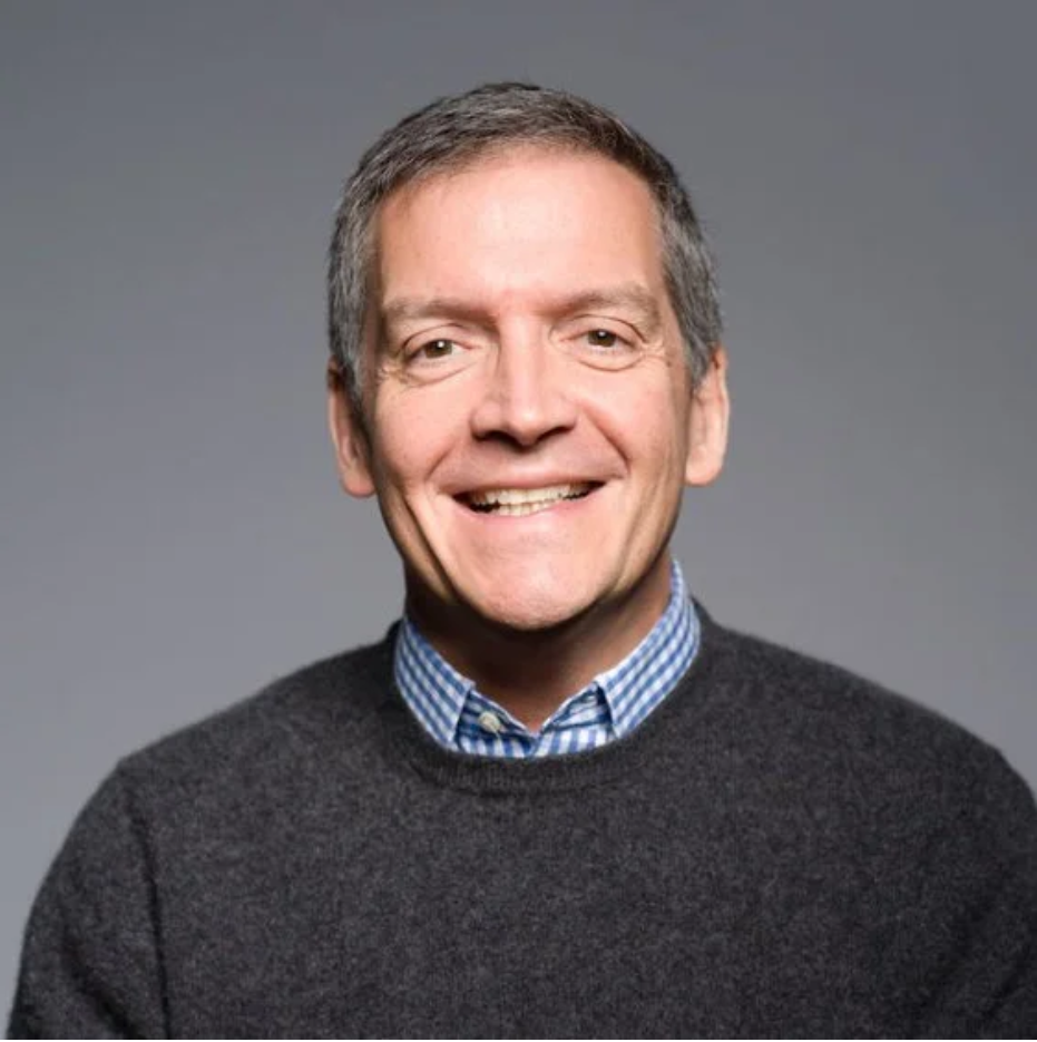 A smiling middle-aged man with short brown hair, wearing a striped blue and white shirt under a dark gray sweater, against a plain gray background.