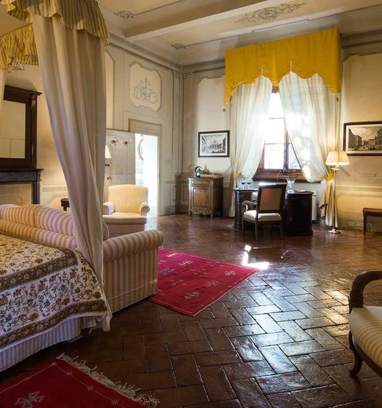 A spacious, well-lit vintage-style bedroom with elegant decor, a canopy bed with striped bedding, a pink area rug, a writing desk with a chair near a window, and classic framed artwork on the walls.