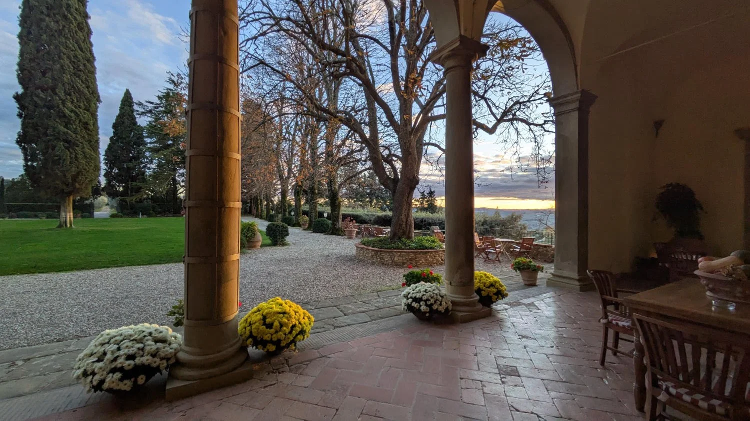 View from a covered patio with columns, looking out onto a landscaped garden with trees, potted flowers, and a sunset in the distance.