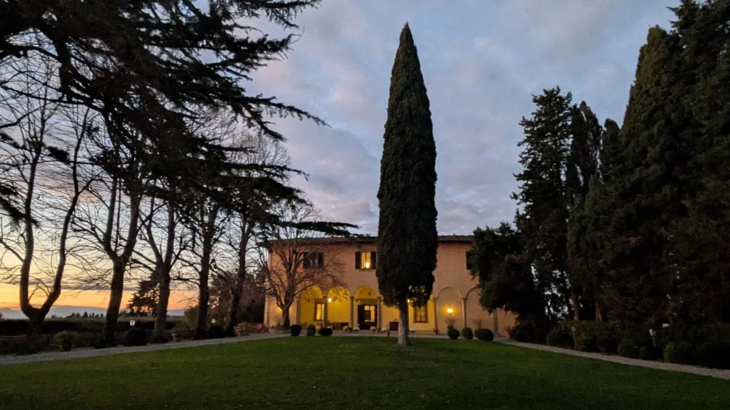 A large house with a pinkish exterior, yellow archways at the front entrance, and windows with lights on, surrounded by tall trees and a well-maintained grassy lawn during dusk or early evening.