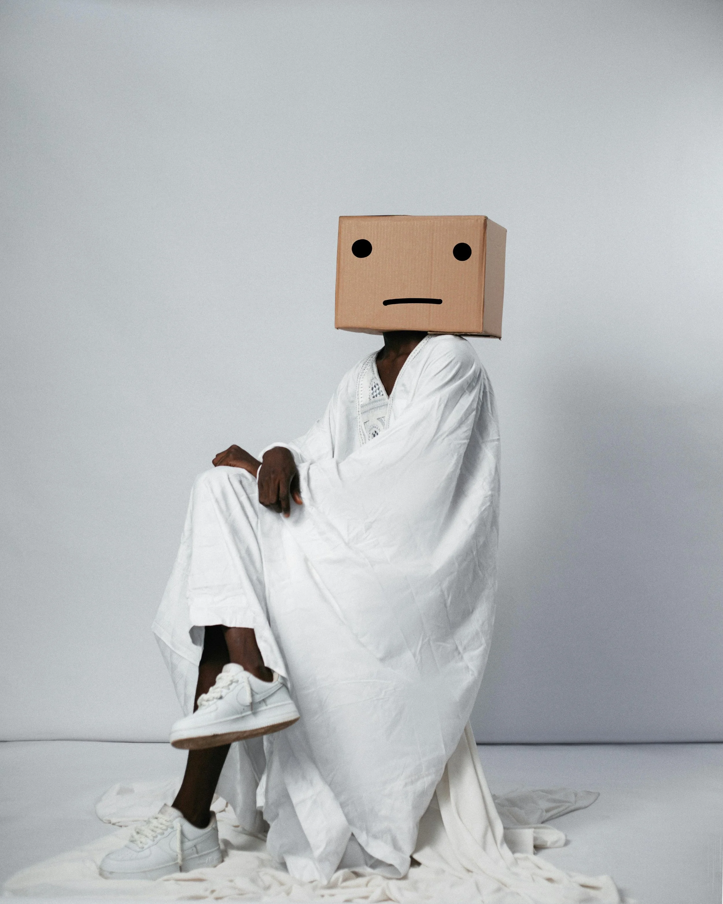 Person with a cardboard box on their head featuring a simple face drawing, sitting on a white cloth-covered surface, dressed in white clothing and sneakers, against a plain light gray background.