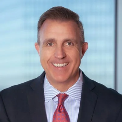 A smiling man wearing a dark suit, light blue shirt, and red tie, standing in front of a window with a cityscape background.