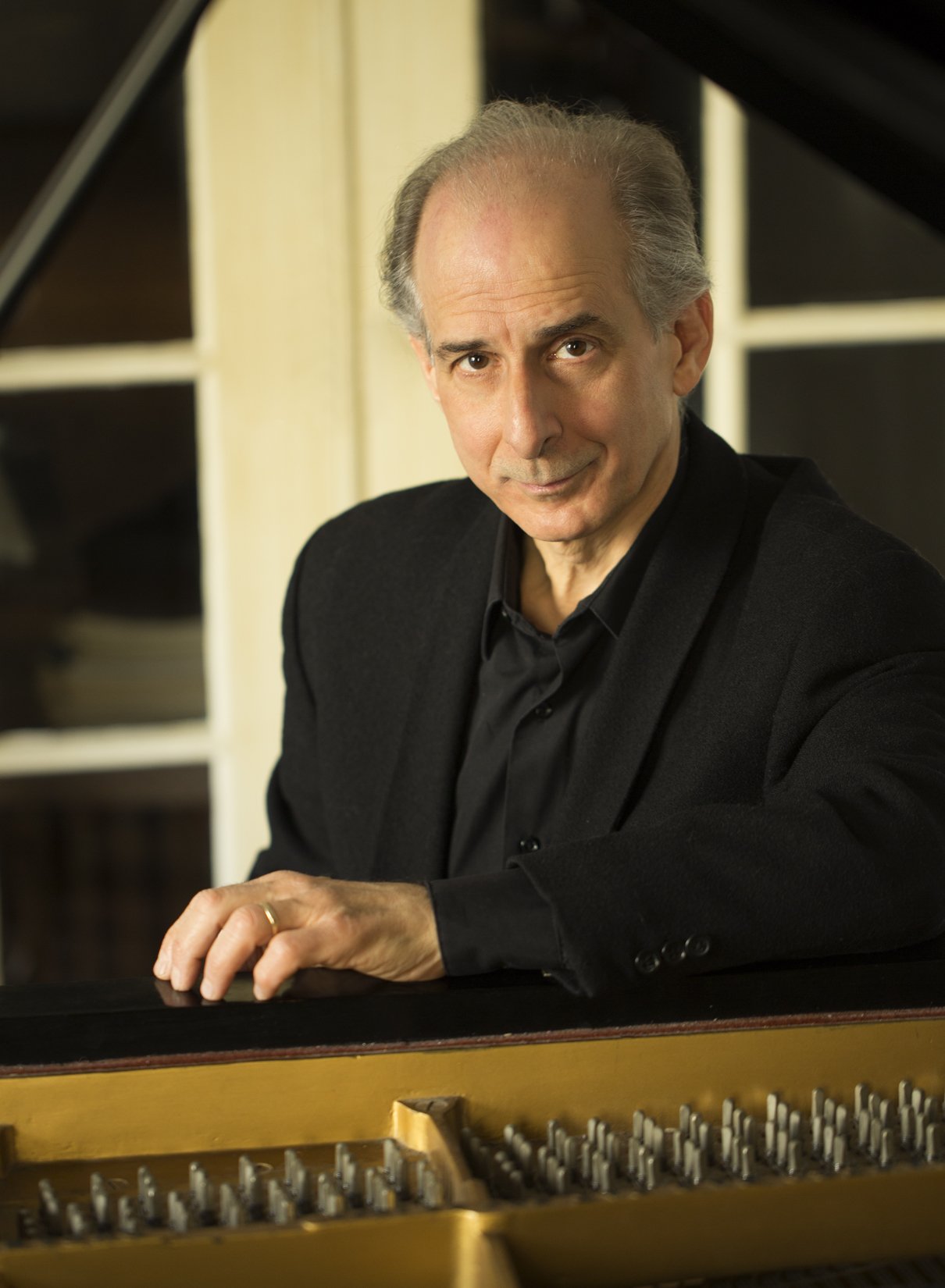 A portrait of an older man with gray hair, wearing a black suit and black shirt, sitting at a grand piano and looking at the camera with a slight smile.