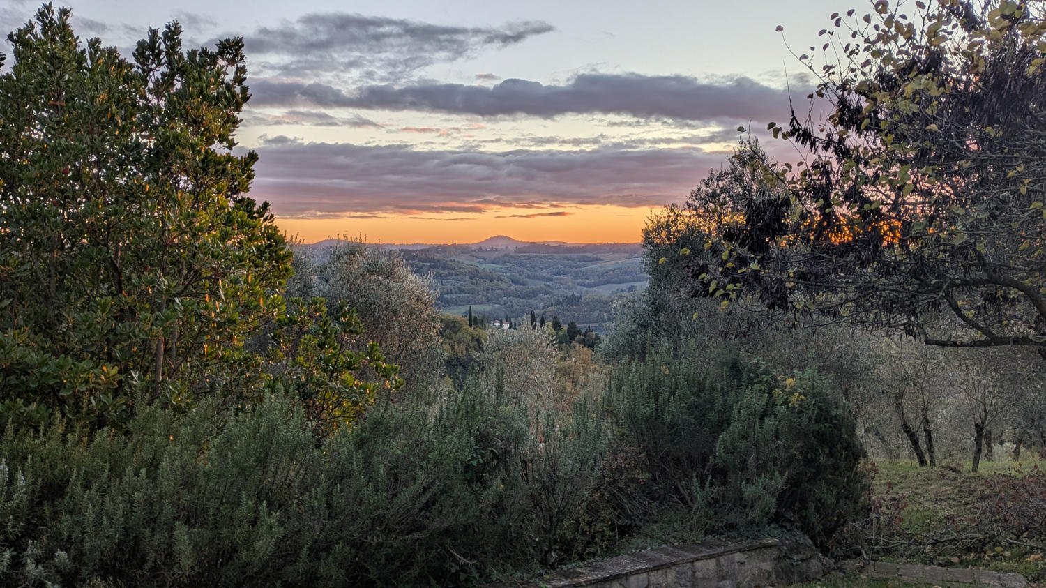 A scenic landscape view during sunset with rolling hills, cloudy sky, and trees in the foreground.