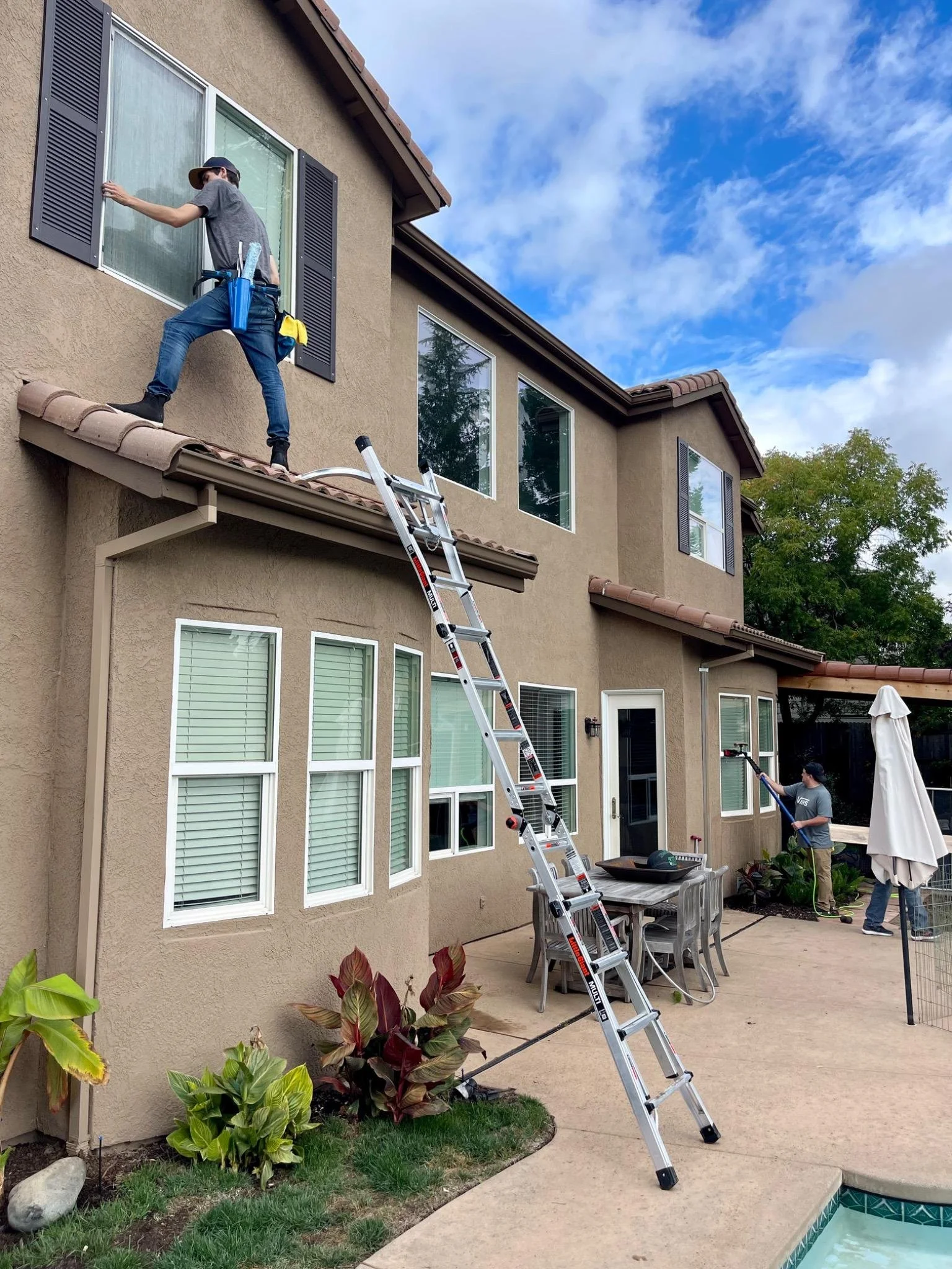 Two workers cleaning windows on a beige stucco house, one on the roof with a ladder and the other on the ground with a pole, during daytime with blue sky and some clouds.