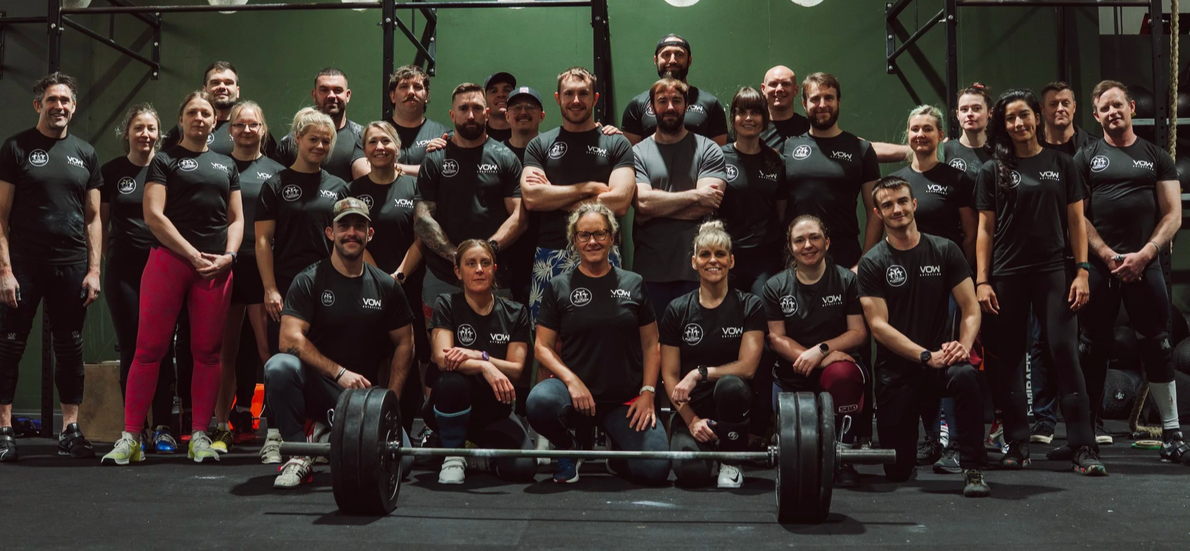 Group of men and women in gym, some wearing black shirts with a white logo that shows The Platform Project, posing for group photo, weightlifting equipment in front.