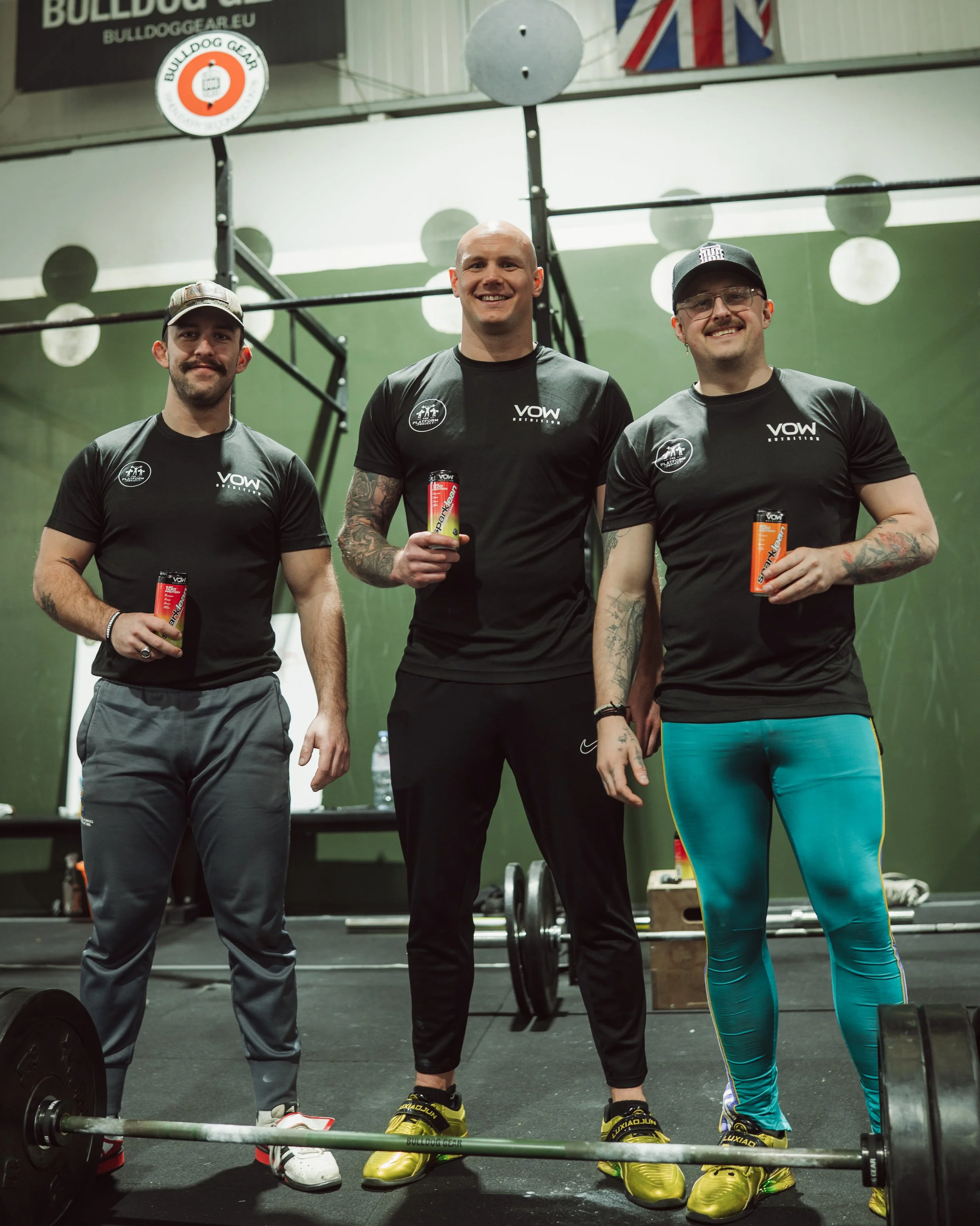 Three men in black athletic shirts with 'VOW' logos, standing in a gym, each holding a red energy bar. They are smiling, with one man in the center holding a protein shake, and all appear to have tattoos. Gym equipment and weights are visible in the background.