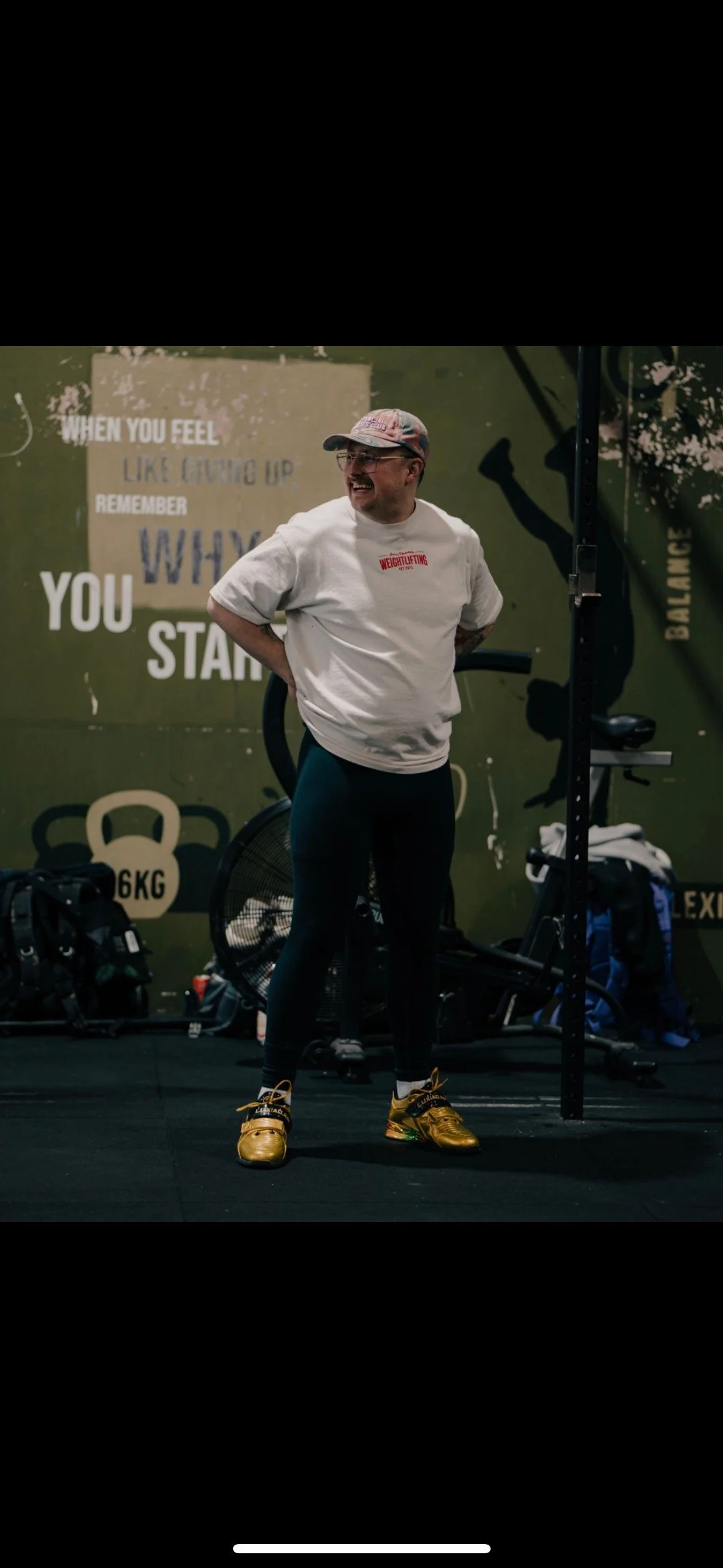 Man in white T-shirt and gold weightlifting shoes standing in gym with motivational wall posters and fitness equipment behind him.