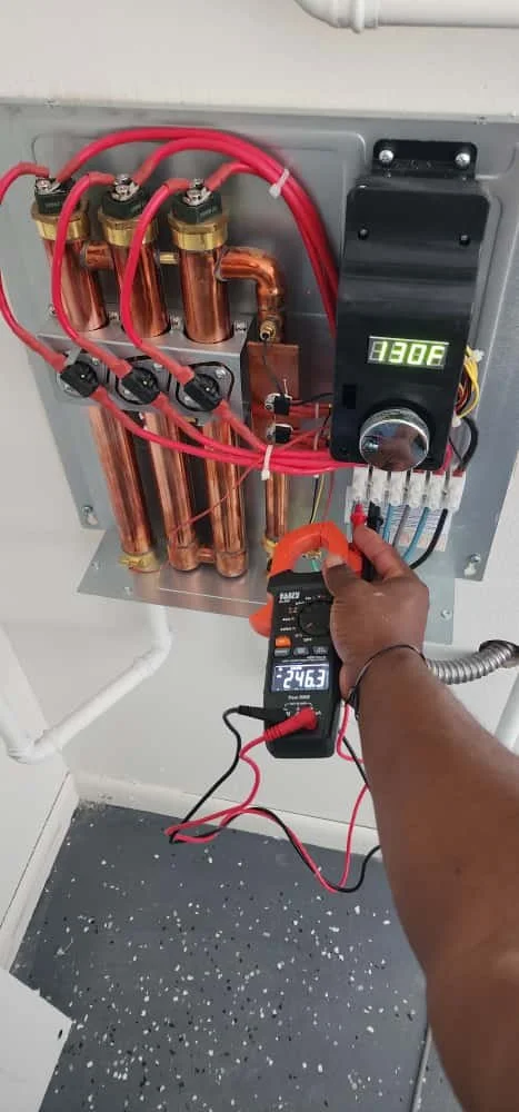 Technician is testing a tankless water heater. The panel contains copper coils, red wiring, and a black control unit displaying 130°F.