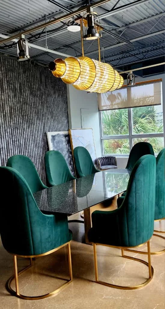 Modern dining room with six green velvet chairs around a black granite table, gold accents, large window with greenery outside, contemporary hanging light fixture, and artwork leaning against the wall.