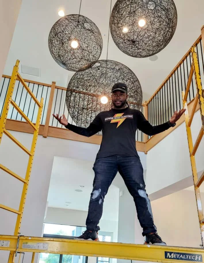 South Florida electrician standing on a yellow scaffolding inside a modern home, with intricate black wire pendant lights hanging from the ceiling above him.