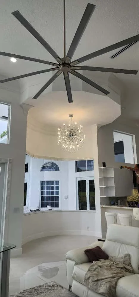 Interior view of a living room with a large, modern ceiling fan, a chandelier, white walls, and a white sectional sofa with throw pillows and a blanket.
