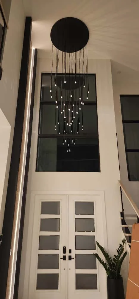 Modern chandelier with pendant lights hanging from a black circular fixture in a high-ceiling entryway, with large windows, white doors, and a potted plant.