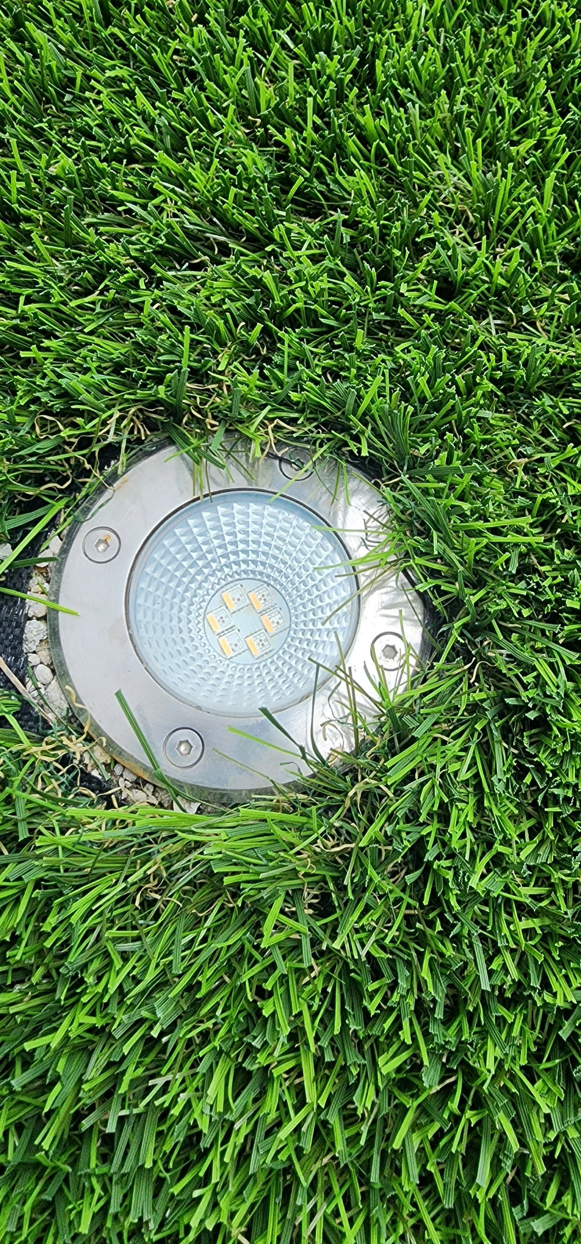 A round metal outdoor LED light embedded in the ground, surrounded by lush green grass.