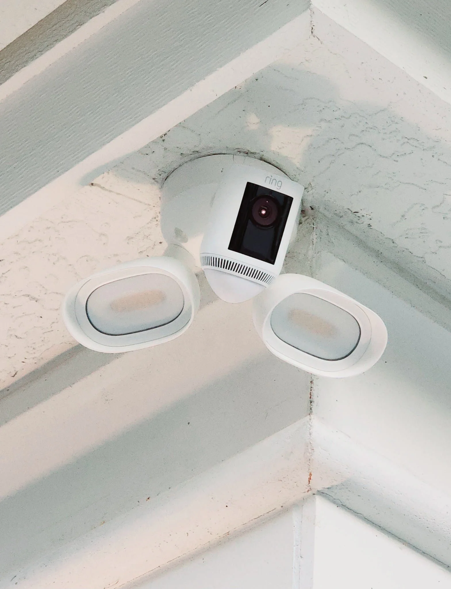 White security camera that was installed by power bolt electric mounted on a ceiling with two white light fixtures on either side.