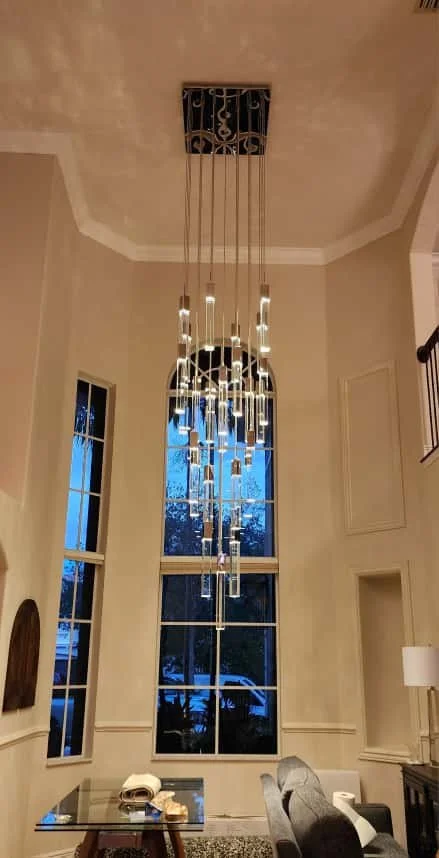 Modern chandelier just installed by an Electrician Boca Raton with multiple hanging glass lights hanging in front of tall windows in a high-ceiling living room.