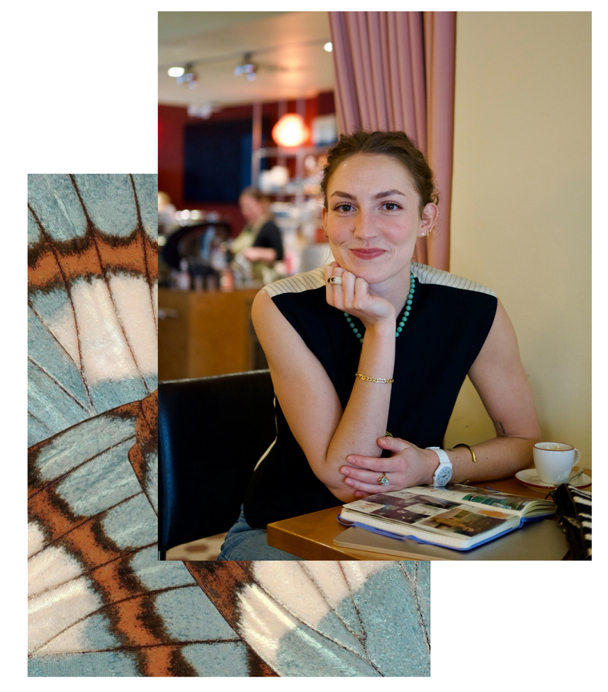 A young woman sitting at a table in a cafe or restaurant, resting her chin on her hand and smiling. She has a necklace, bracelets, and rings, with an open magazine, a cup, and a purse on the table.