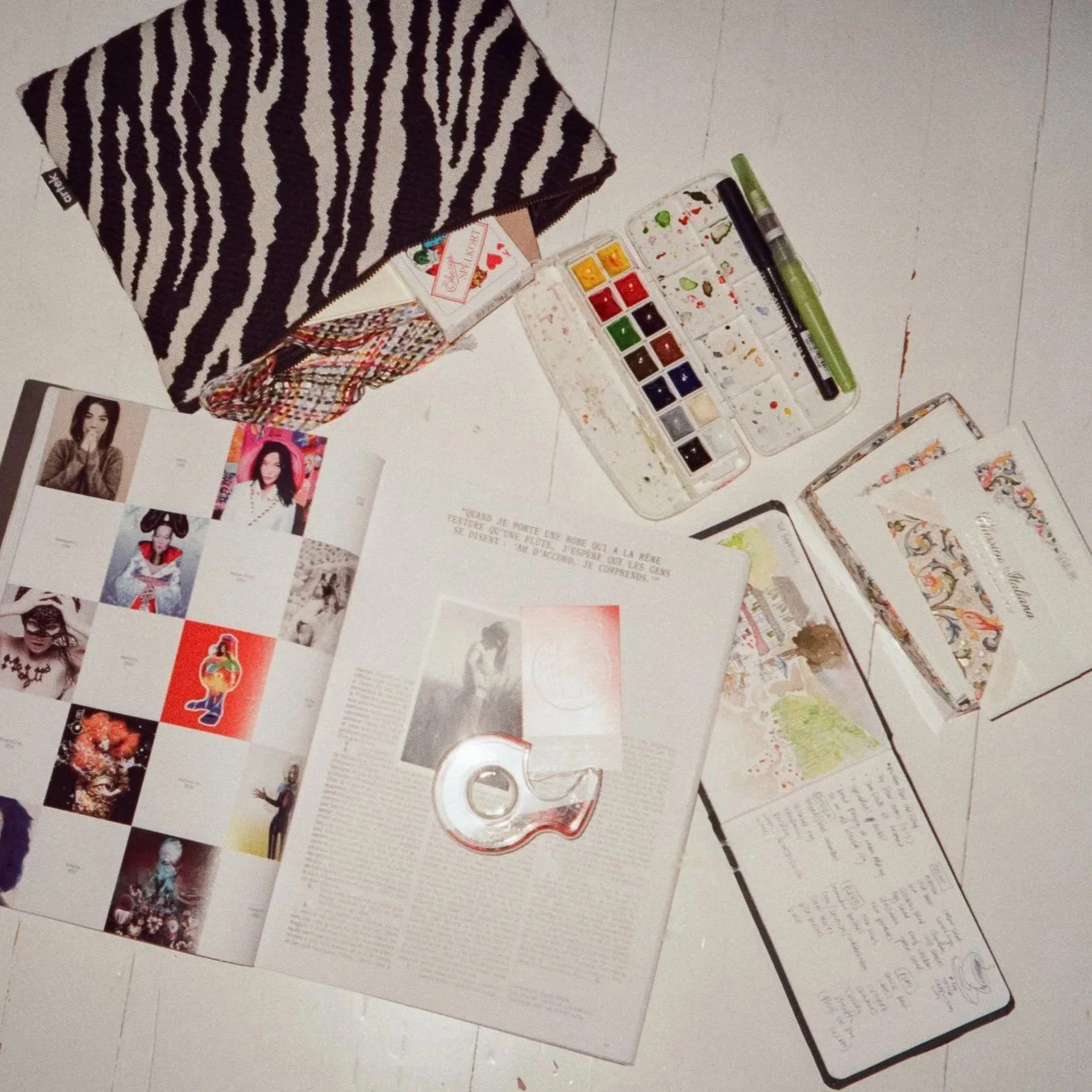 Open magazine with photographs and handwritten notes on a white surface, watercolor paint set, notebooks, a pen, and a zebra print pouch.