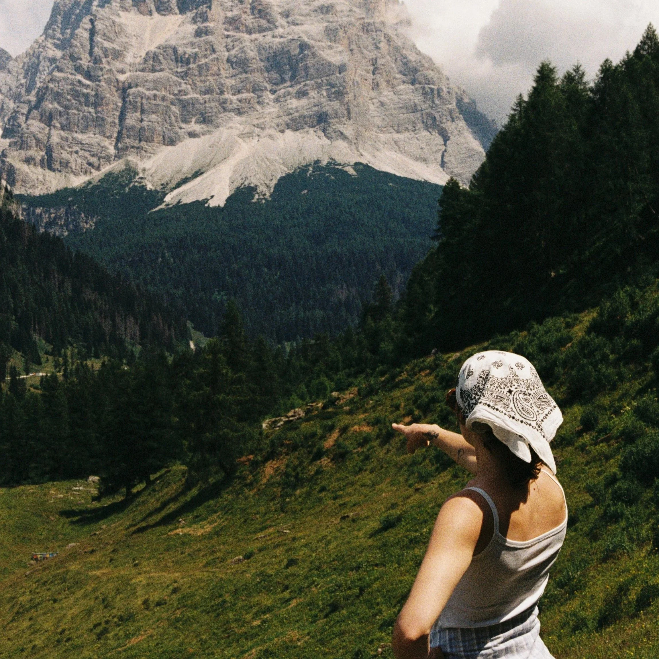 A woman wearing a white bandana and tank top pointing towards a mountain landscape with snow-capped peaks, green forest, and a grassy hillside.