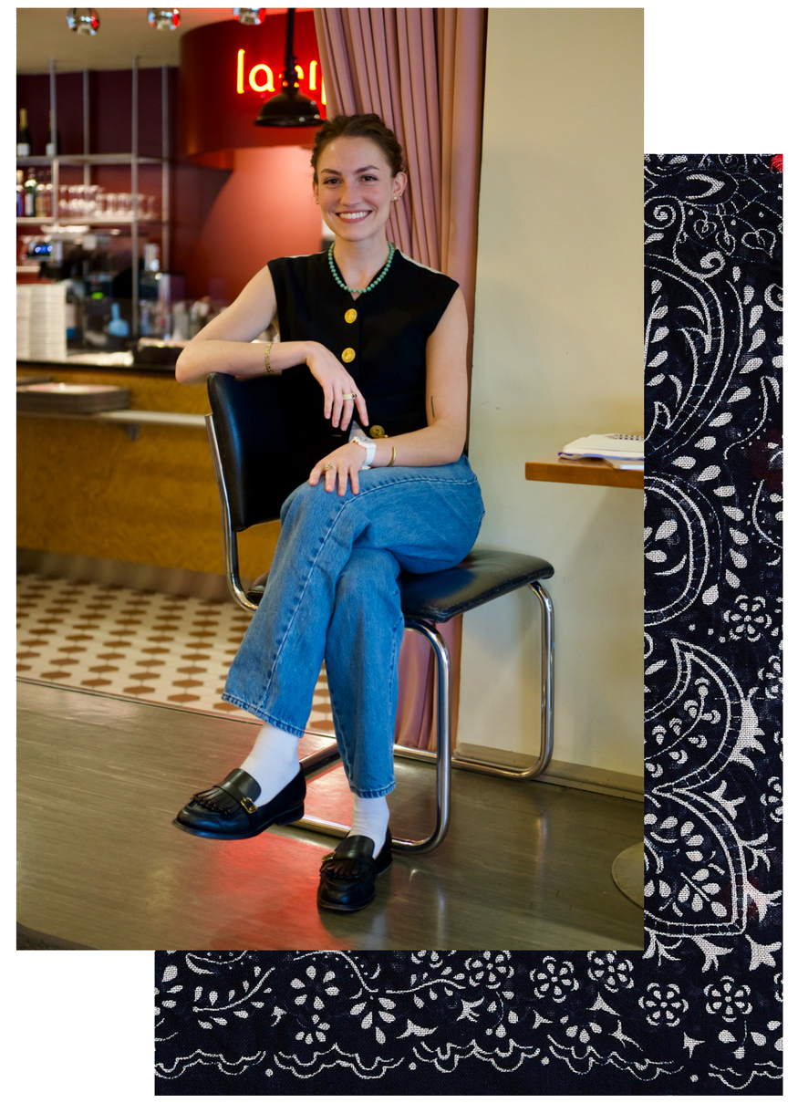 A young woman smiling and sitting on a black chair at a cafe or restaurant. She is wearing a black sleeveless top, blue jeans, white socks, and black loafers, with jewelry including a pearl necklace and bracelets. Behind her is a counter with glasses and utensils, and a red sign with illuminated letters.