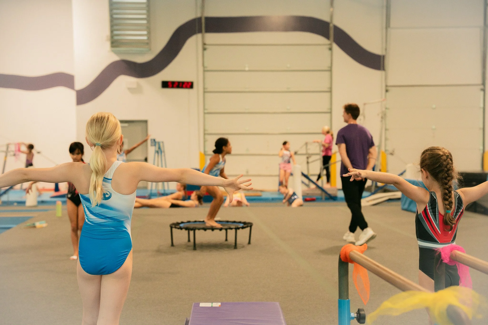 Young girls practicing gymnastics in a training gym, some on the floor, one on a mini trampoline, a coach observing, with a large wall and gym equipment in the background.