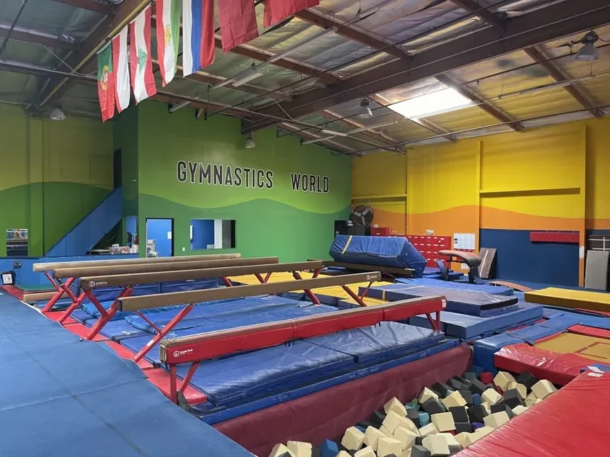 Indoor gymnastics facility with various mats, trampolines, foam pits, and balance beams, with a painted wall reading 'GYMNASTICS WORLD' in the background.