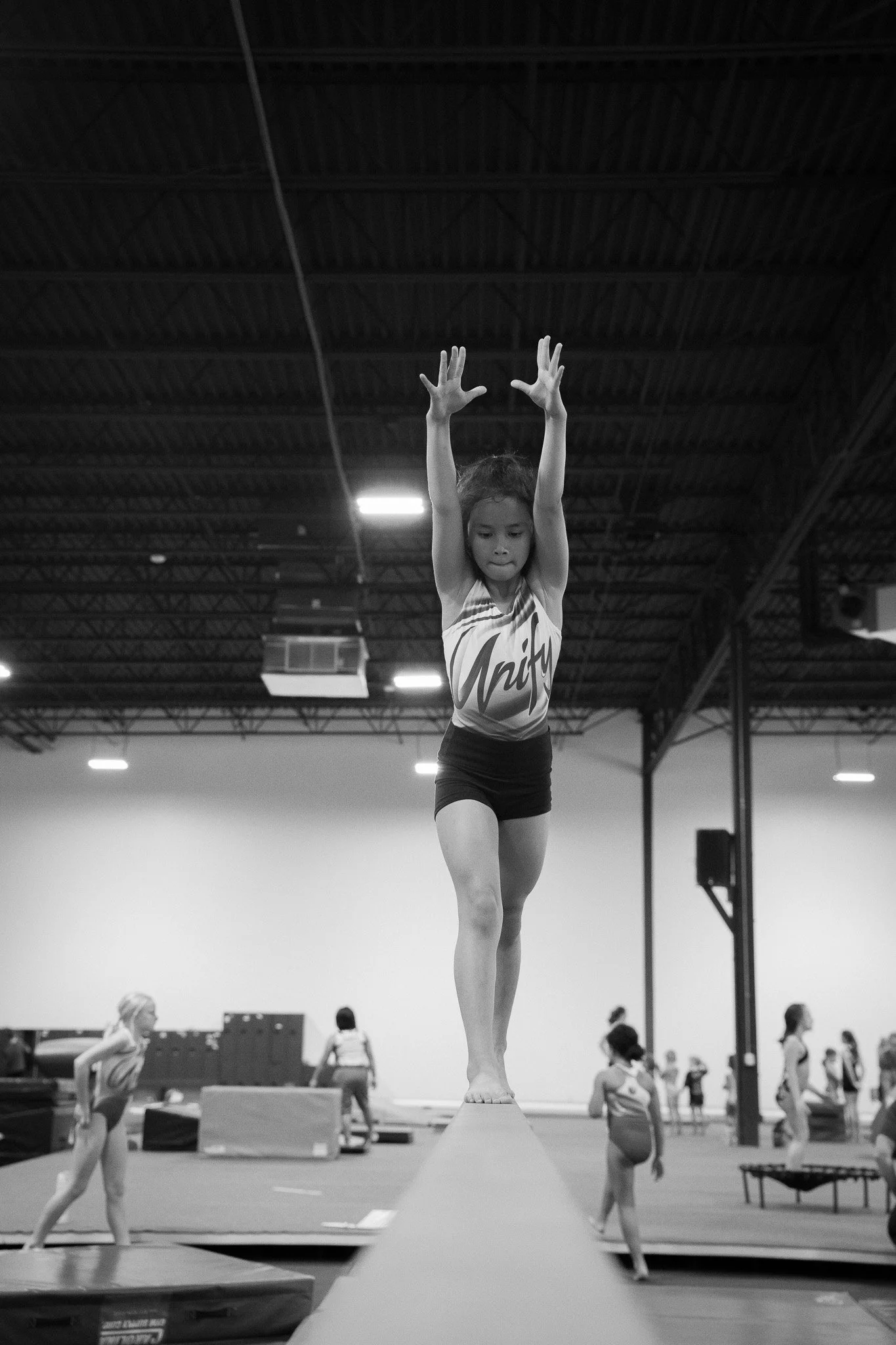 A young girl practicing gymnastics on a balance beam in a gymnasium, with other children in the background practicing on different equipment.